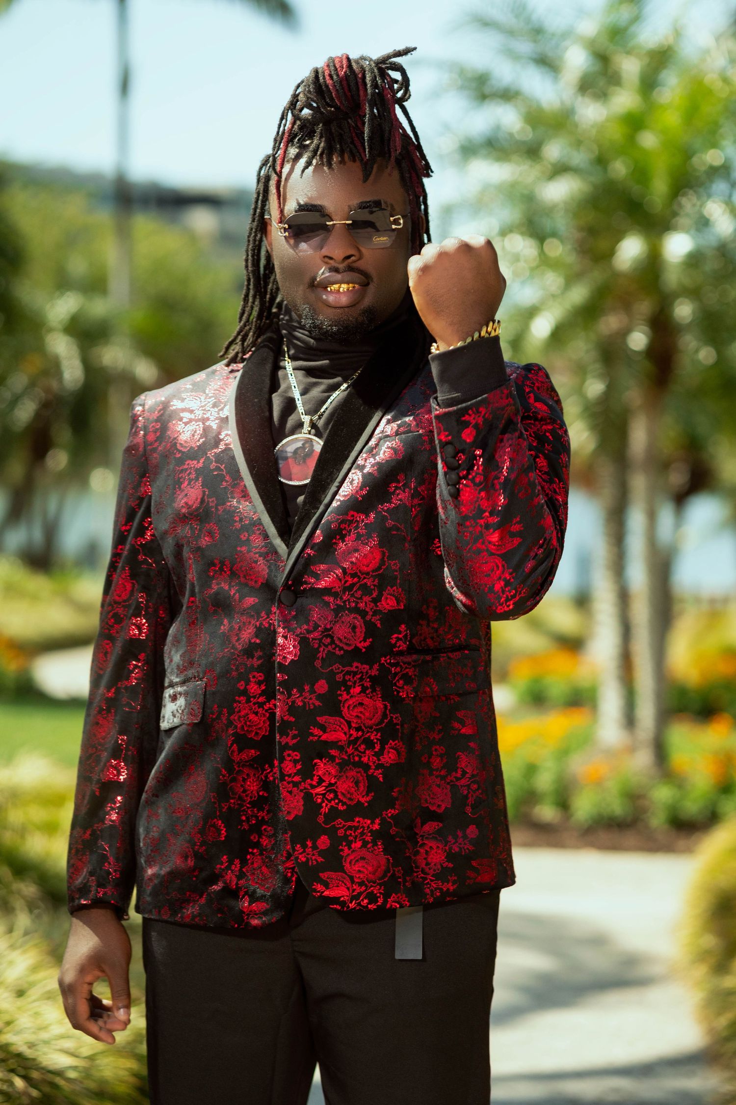 Stylish man in red floral blazer raises fist wearing dreadlocks and gold jewelry outdoors.