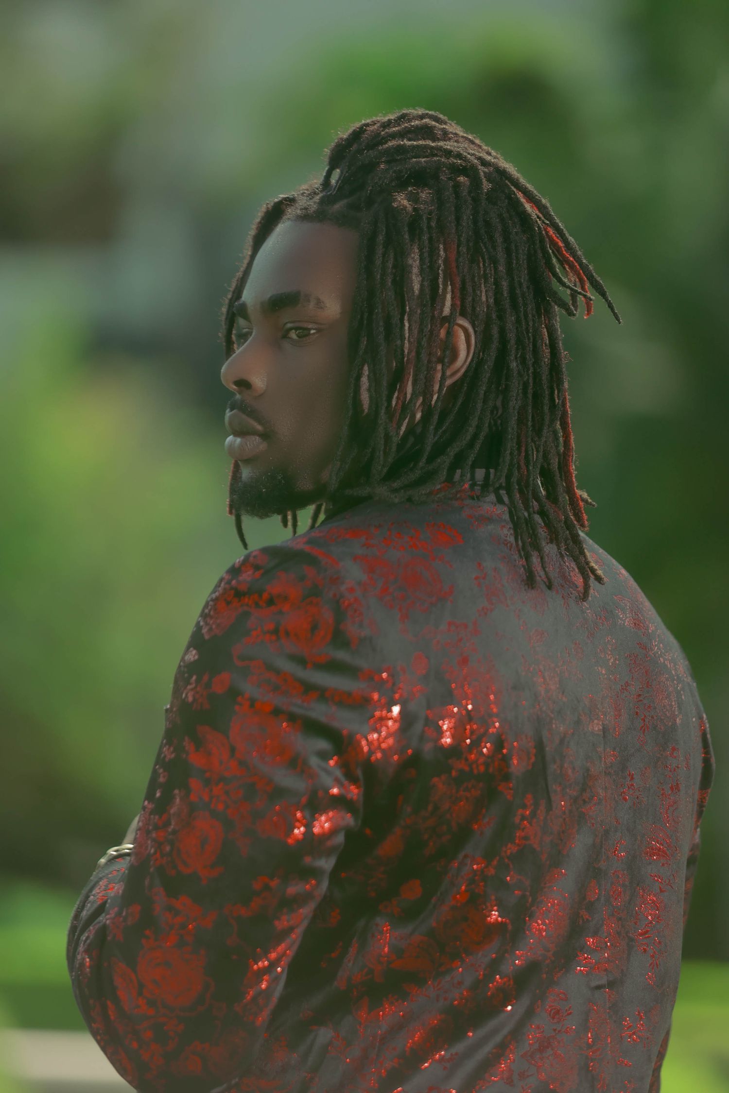 Man with long dreadlocks poses in red sequin floral blazer with arms crossed in green setting.
