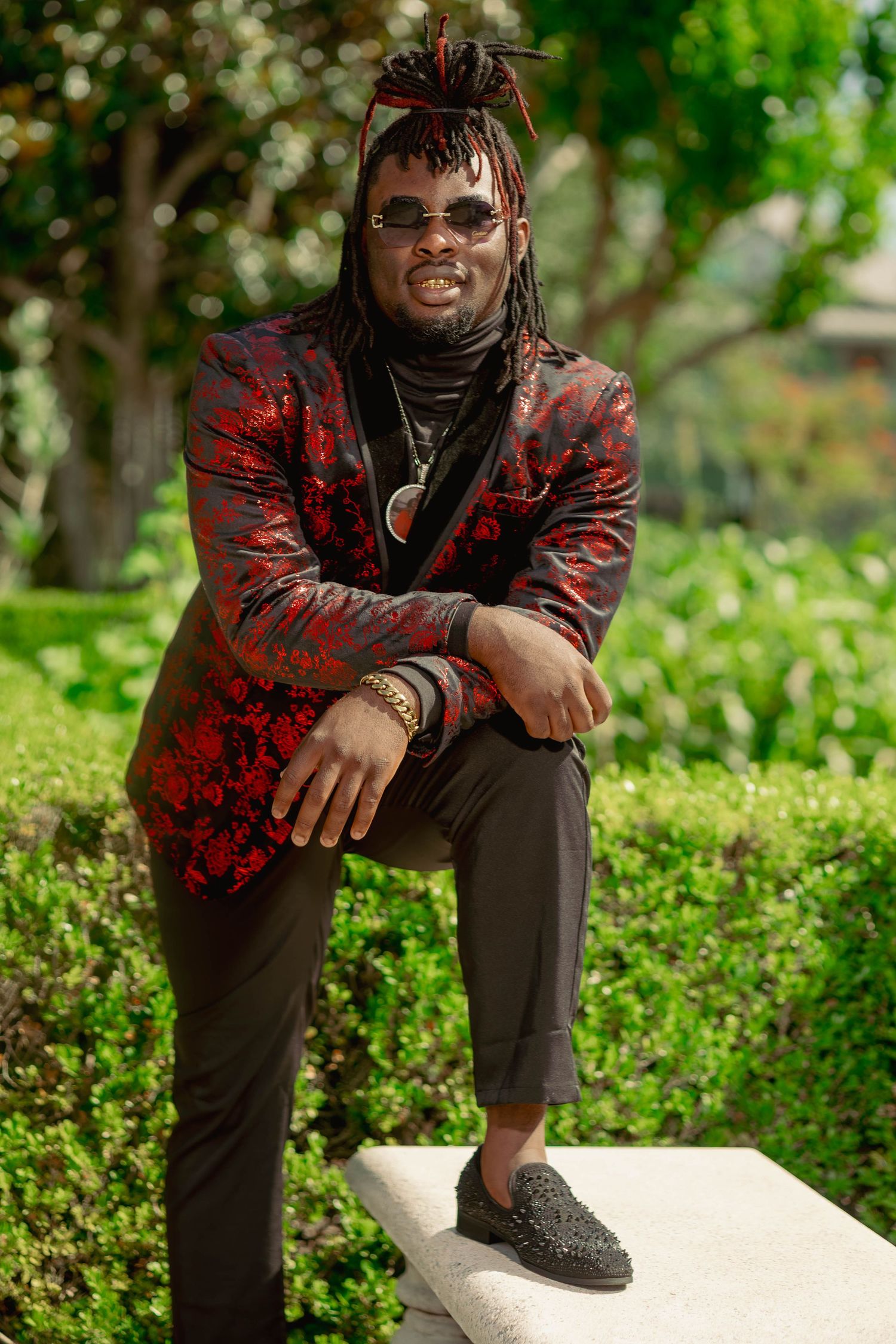 Fashionable man in red floral blazer sits on ledge in lush garden wearing sunglasses and jewelry.