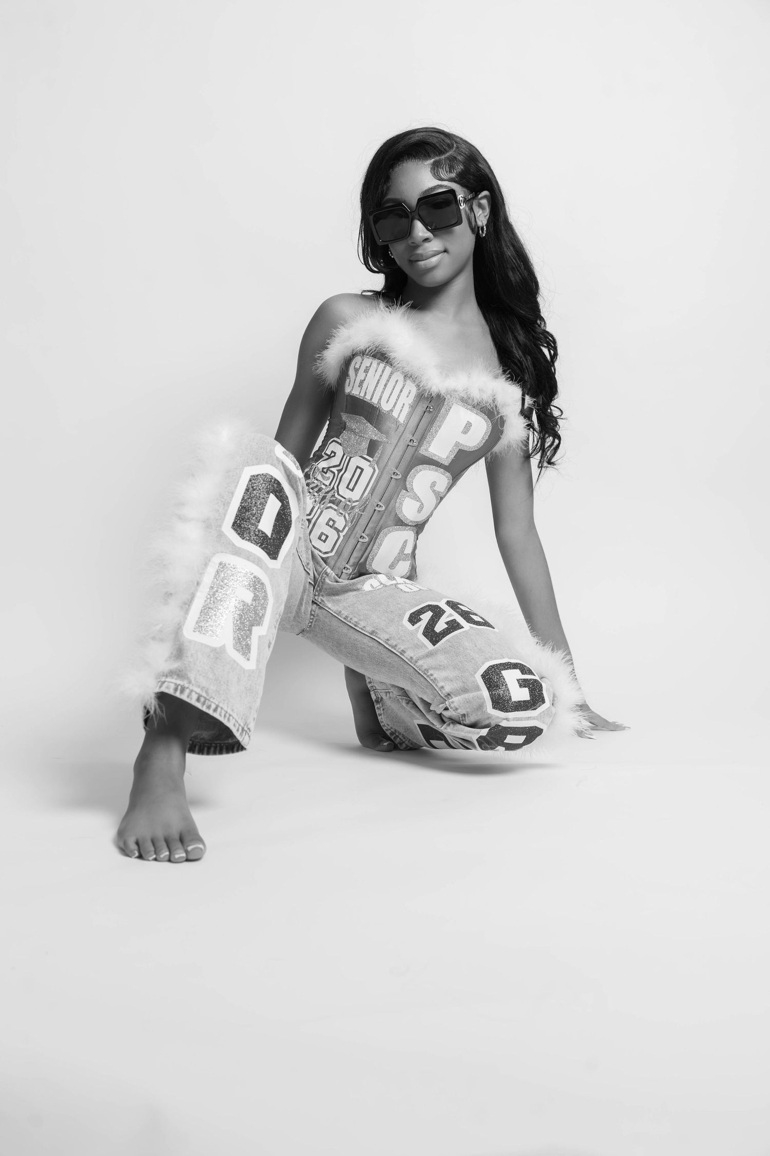 Black and white photo of young woman in designer jeans and sunglasses sitting on floor.