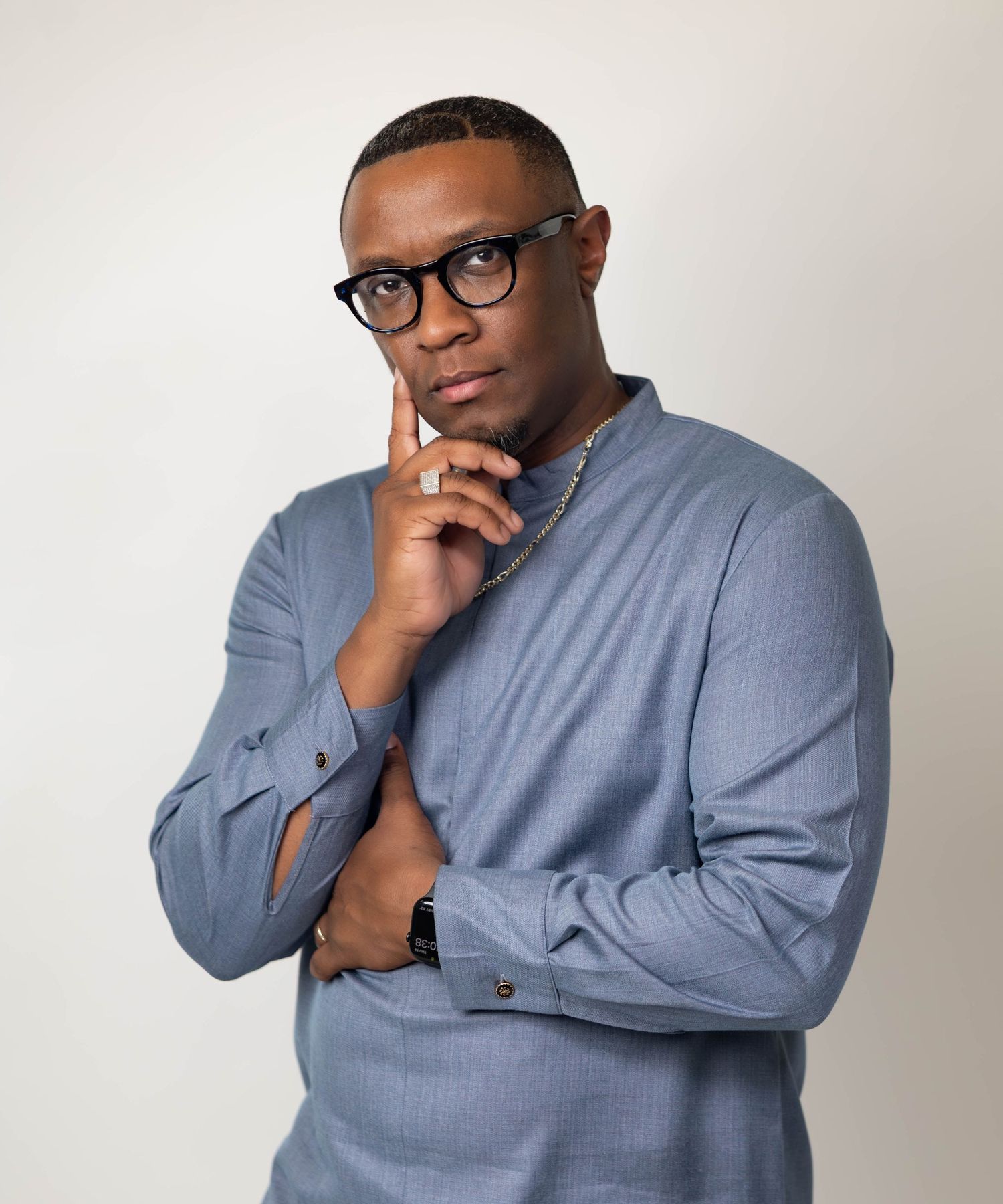 Thoughtful Black man in blue shirt with glasses posing with hand on chin against light background.