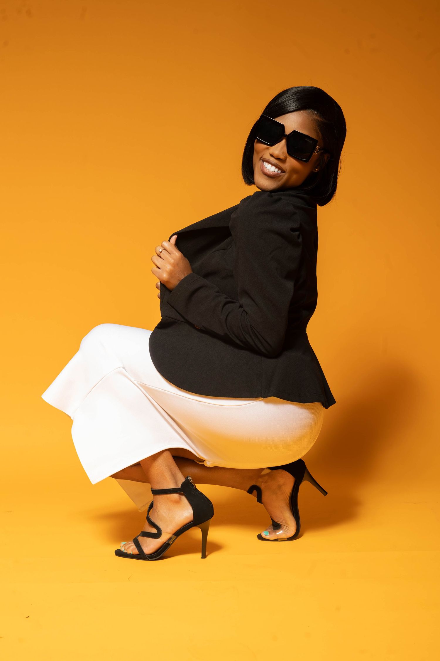 Stylish Black woman in black blazer and white pants crouching with sunglasses on yellow background.