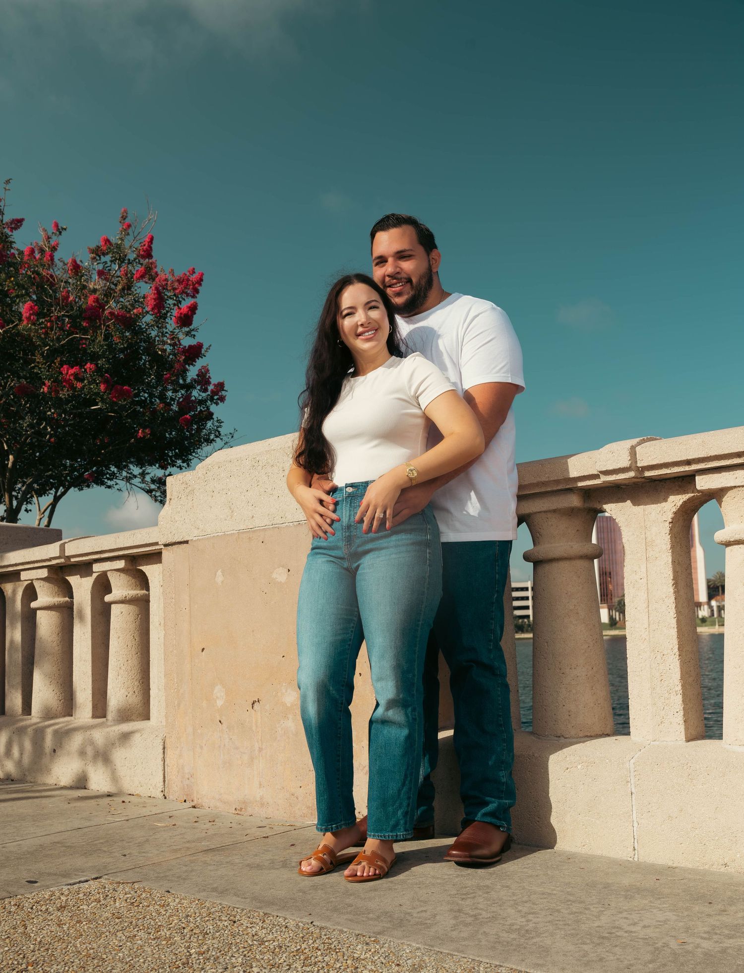 Smiling couple in matching teal jeans posing together on a waterfront promenade.