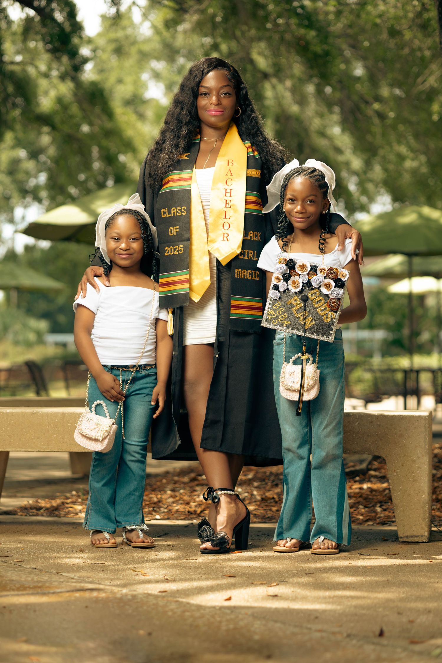 Graduate in Class of 2025 stole poses with two young girls in matching outfits at outdoor ceremony.