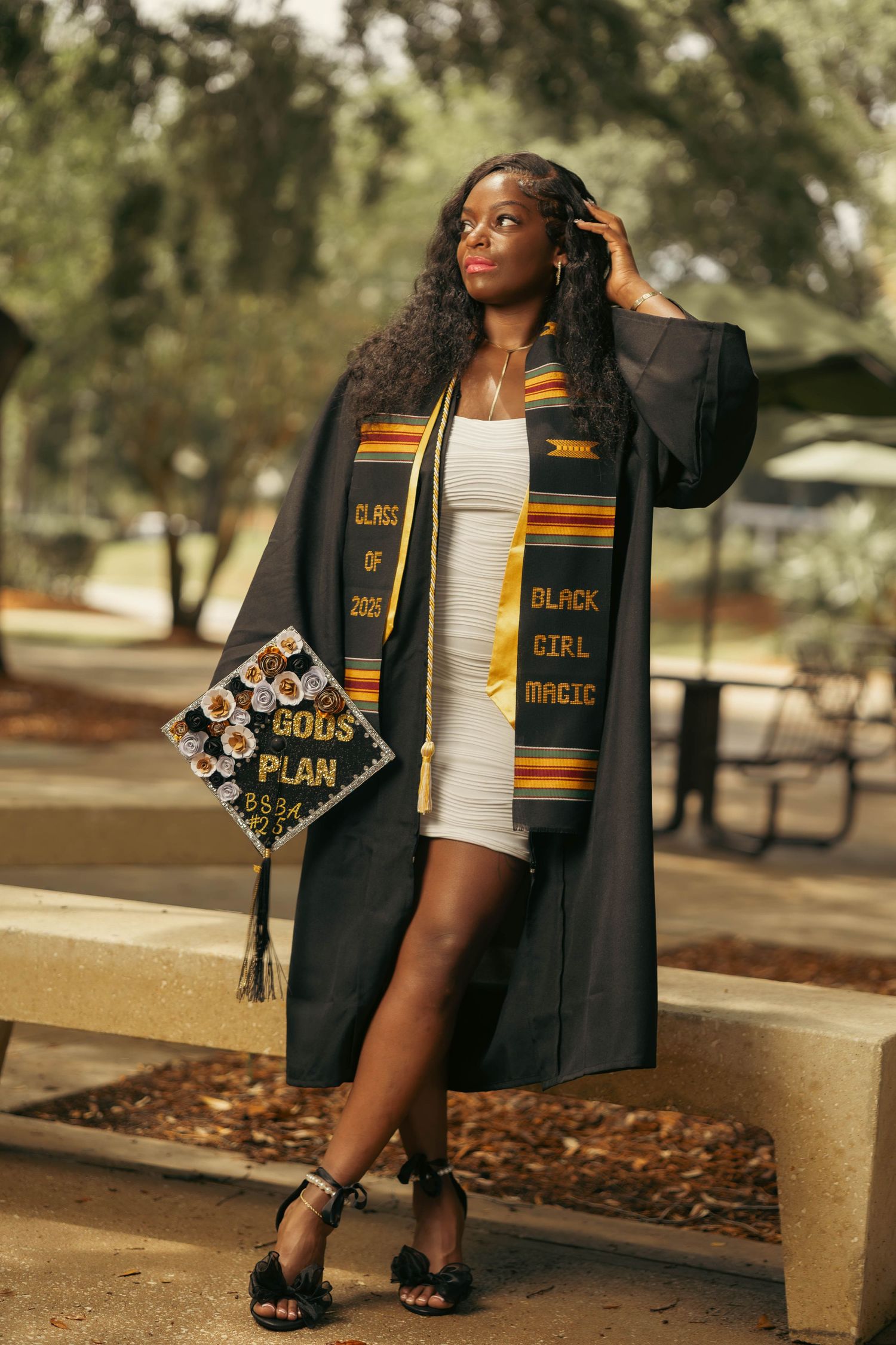 Graduate in black robe with Black Girl Magic kente stole holds decorated Gods Plan cap outdoors.