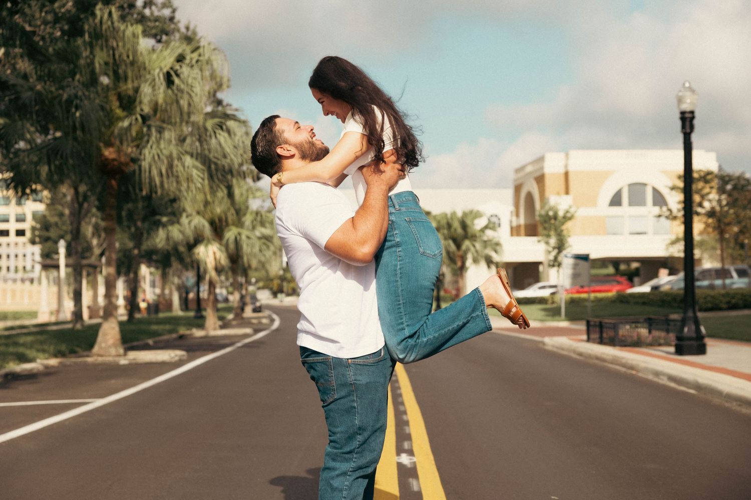 Man lifting woman joyfully in the middle of a palm-tree-lined street outdoors.
