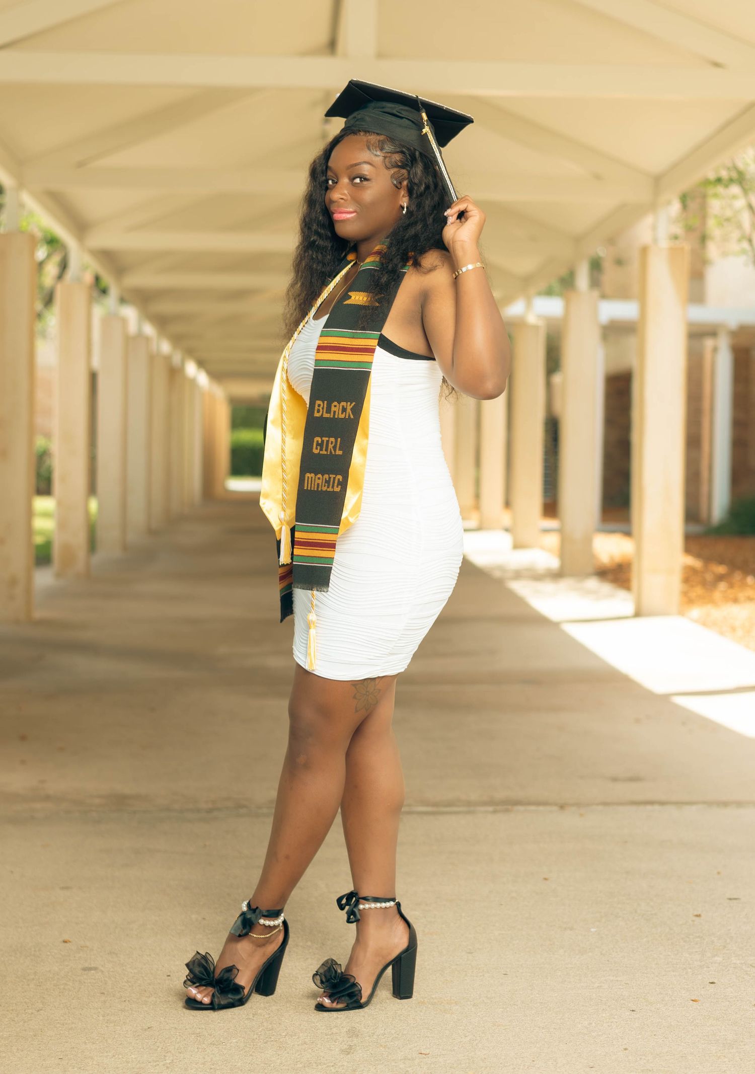 Young Black woman in white dress and graduation cap poses in sunlit corridor with kente stole.