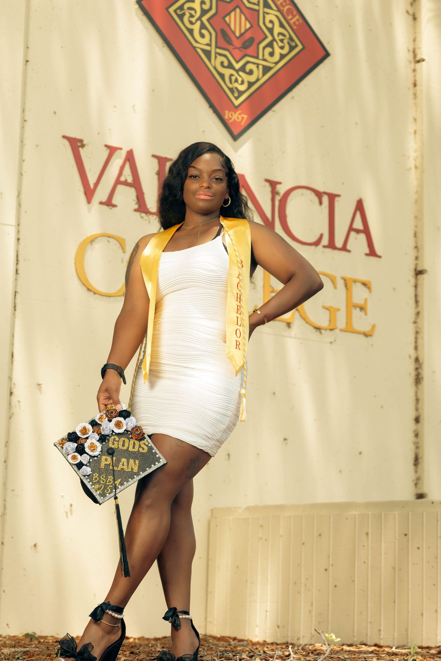 Graduate in white dress holds decorated cap reading Gods Plan before Valencia College sign.