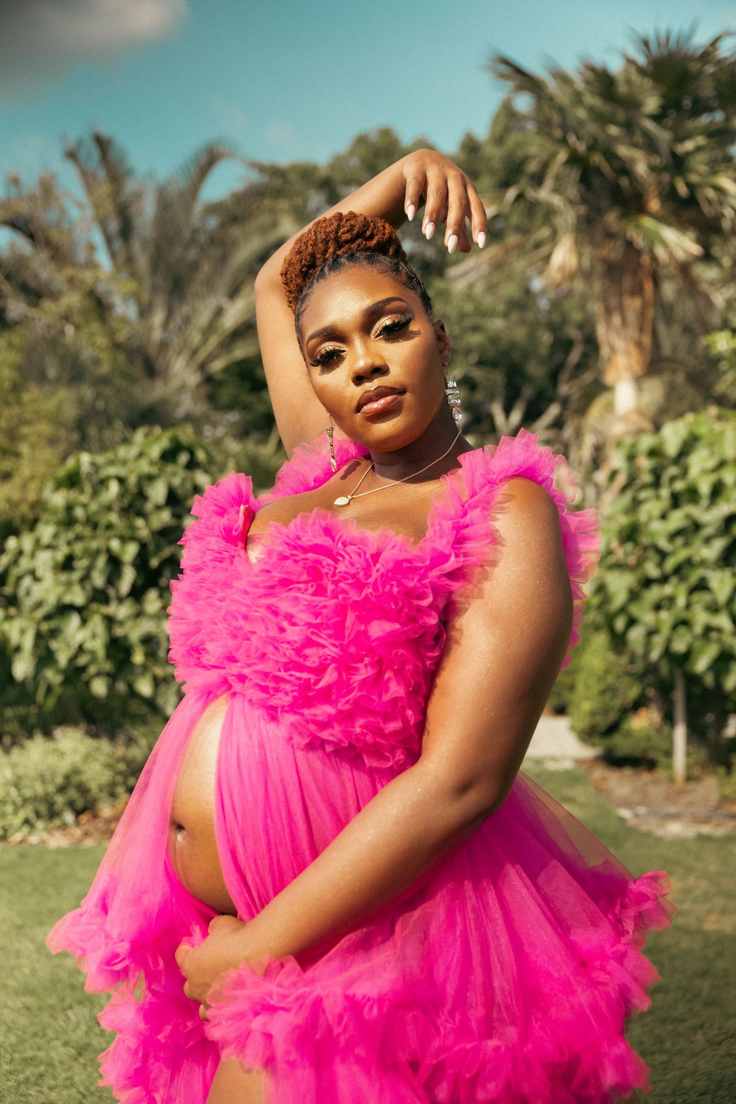Pregnant Black woman in pink ruffled maternity gown posing with arm raised in tropical garden.