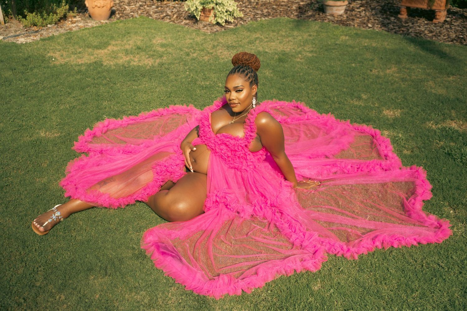 Pregnant Black woman in flowing hot pink ruffled maternity gown seated on lush green grass.
