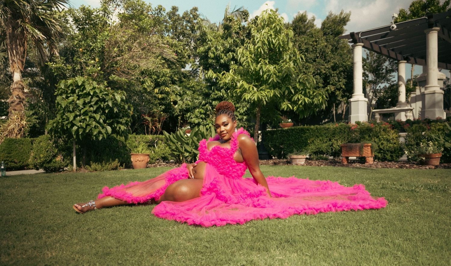 Pregnant Black woman in hot pink ruffled gown sitting gracefully on green lawn in garden.
