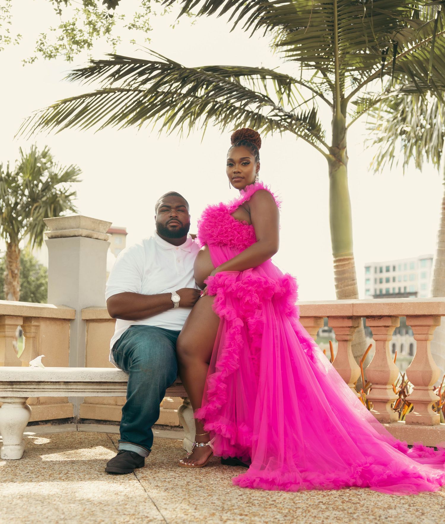 Couple posing outdoors, woman in hot pink ruffle gown and man in white shirt with jeans near palm trees.