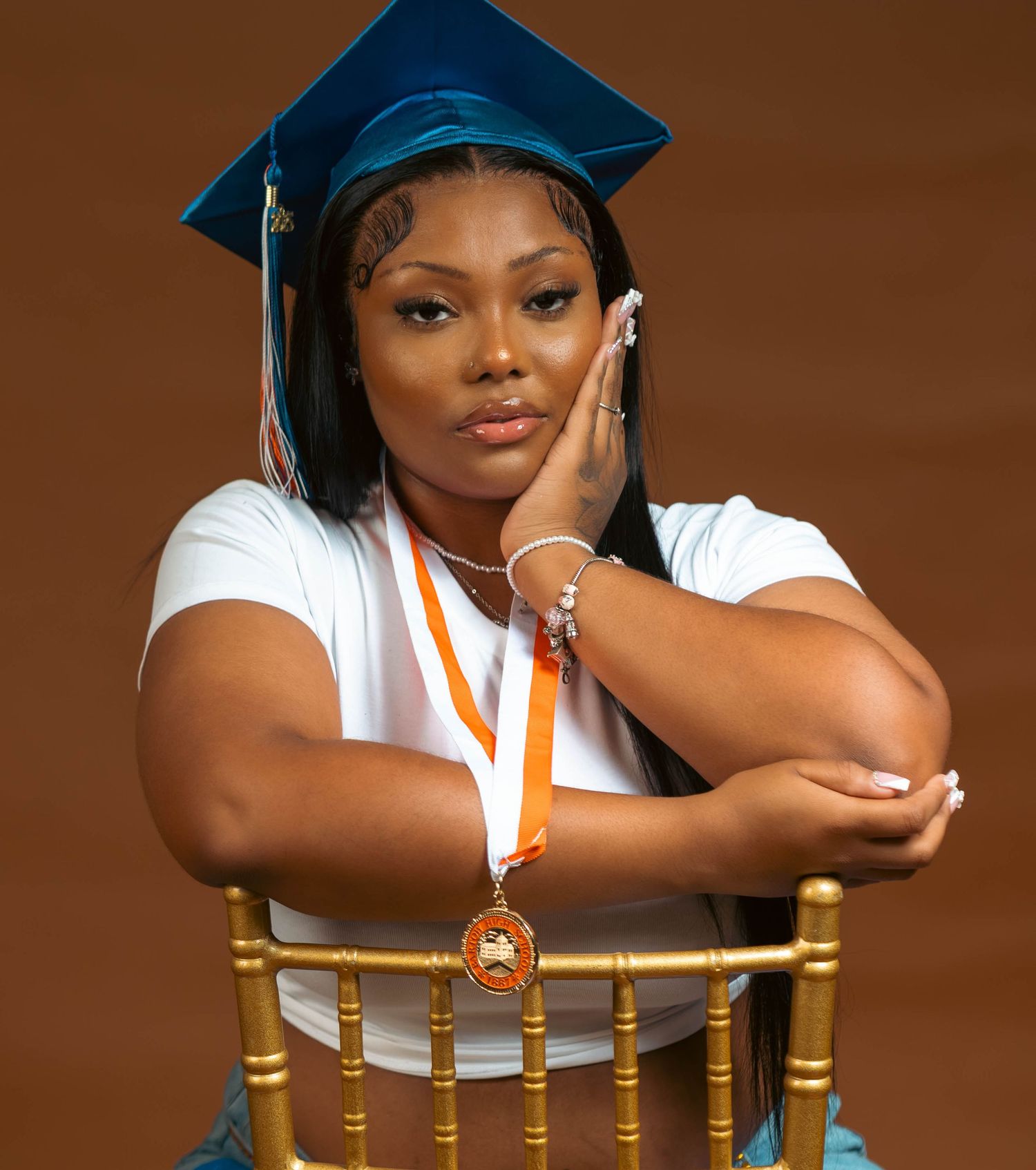 Confident graduate in blue cap with orange medal posing on a gold chair.