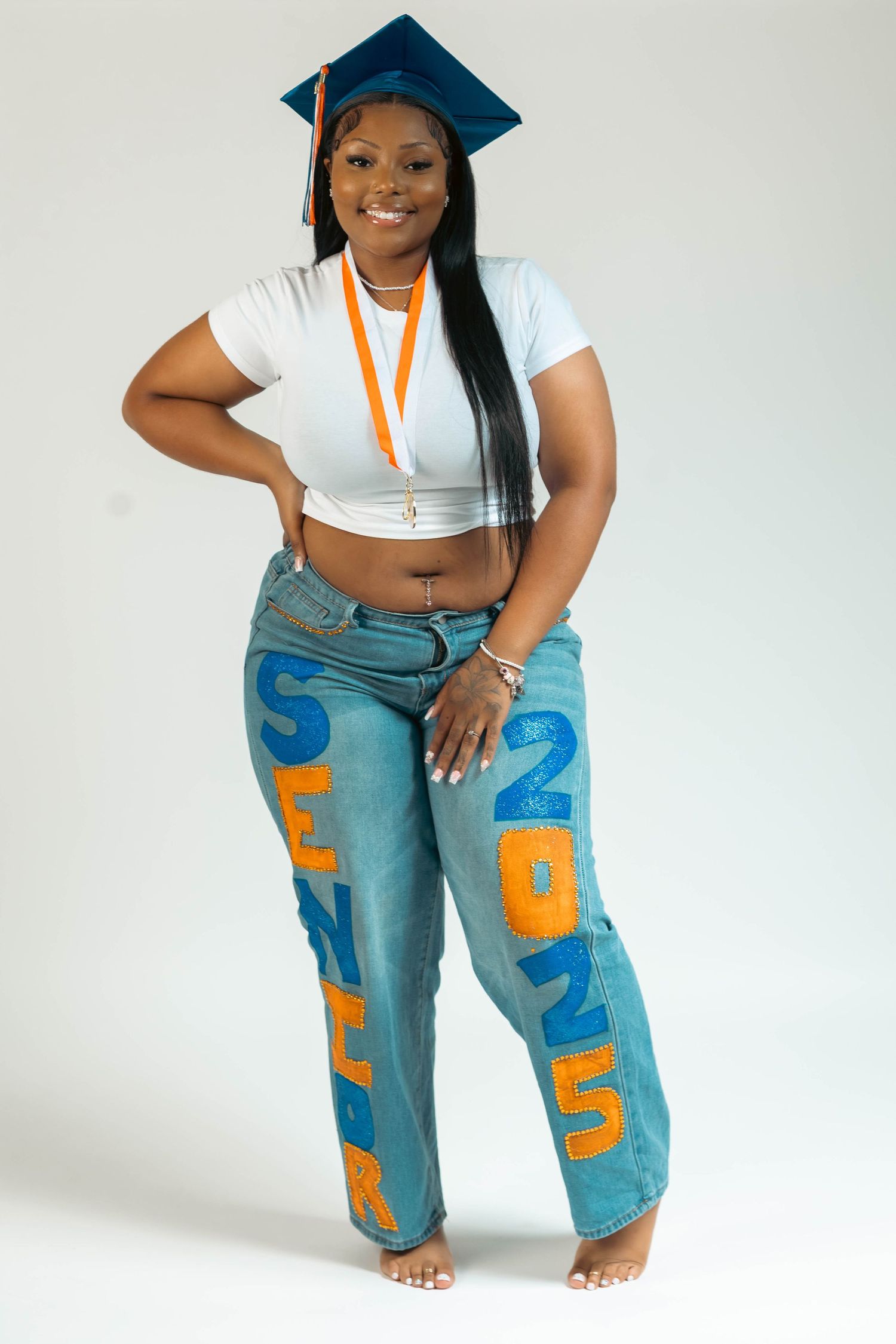 Young Black woman in graduation cap and custom painted jeans with orange and blue letters posing confidently.
