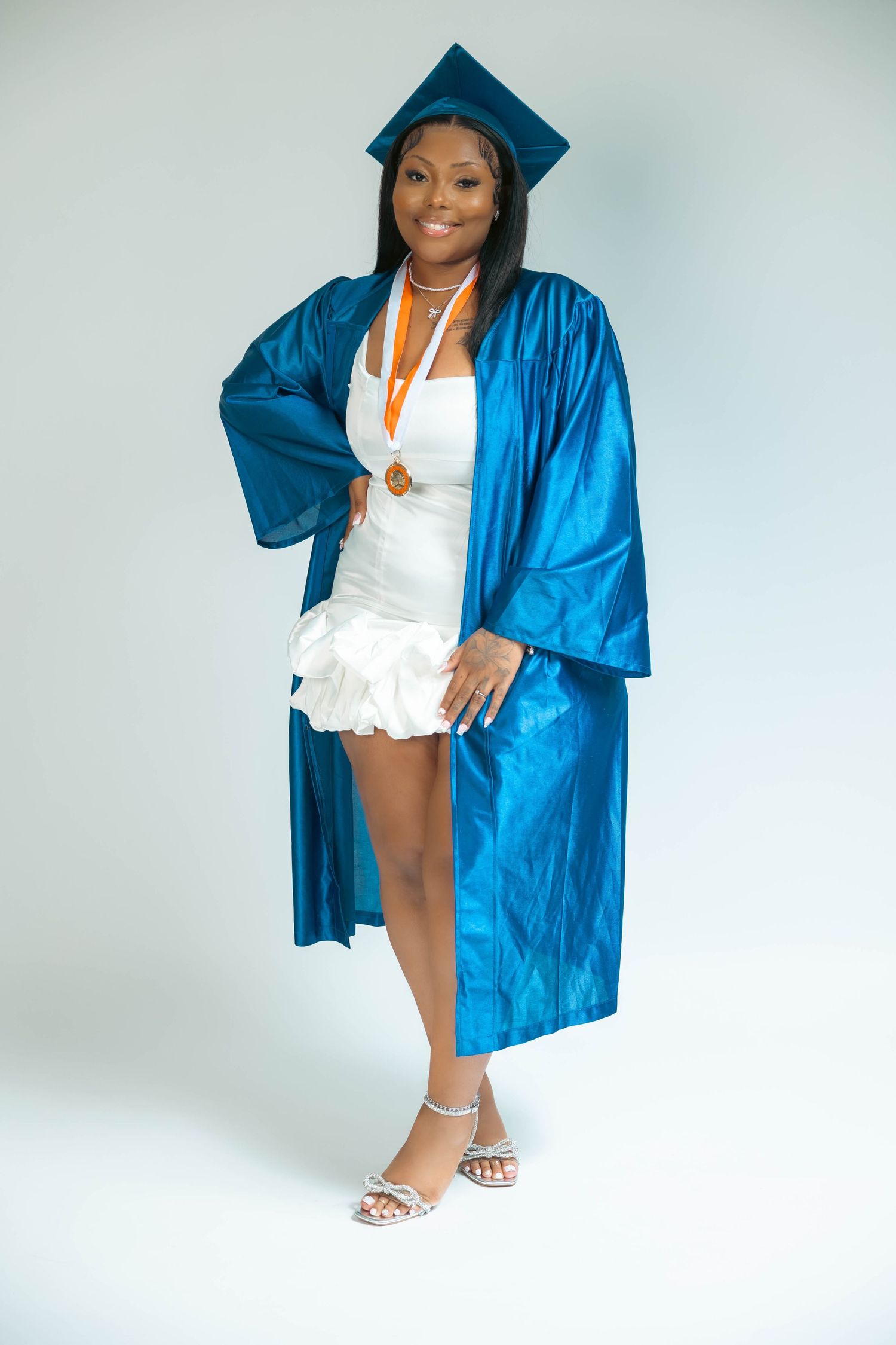 Smiling graduate in blue cap and gown with medal posing in white dress.