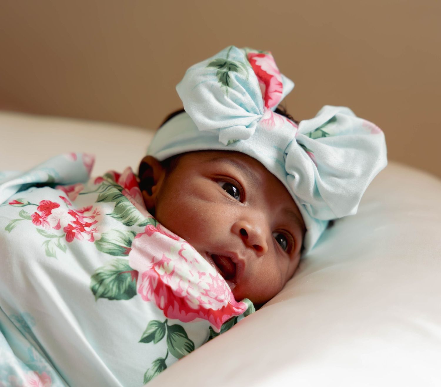 Newborn baby wrapped in floral swaddle with bow headband lying on white surface.