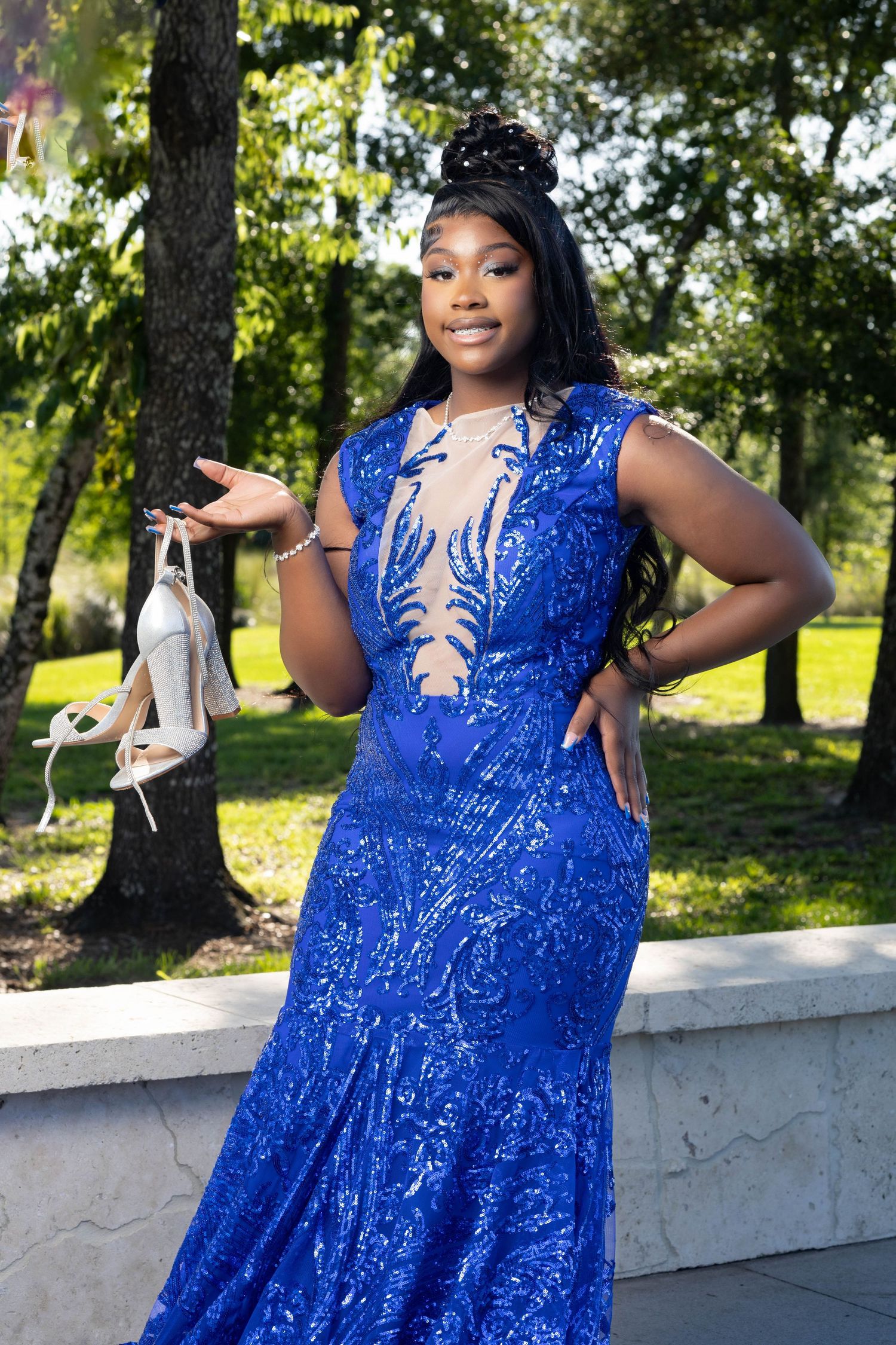 Young Black woman in royal blue sequin prom gown holding heels outdoors in a park.