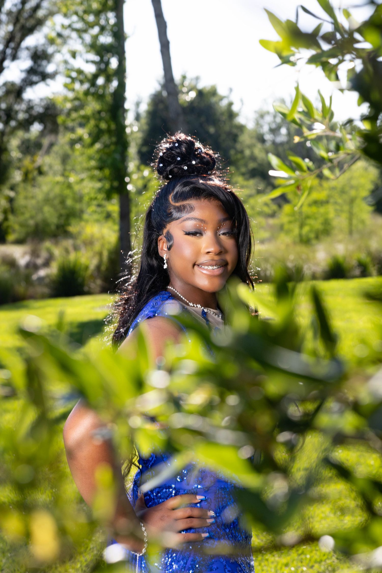 Young Black woman in sparkling blue prom dress smiling among lush green garden foliage.
