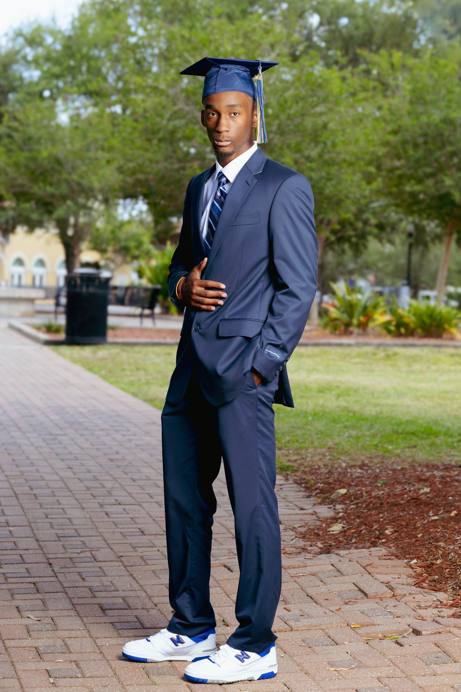 Young male graduate in navy suit and cap posing confidently on an outdoor brick pathway.