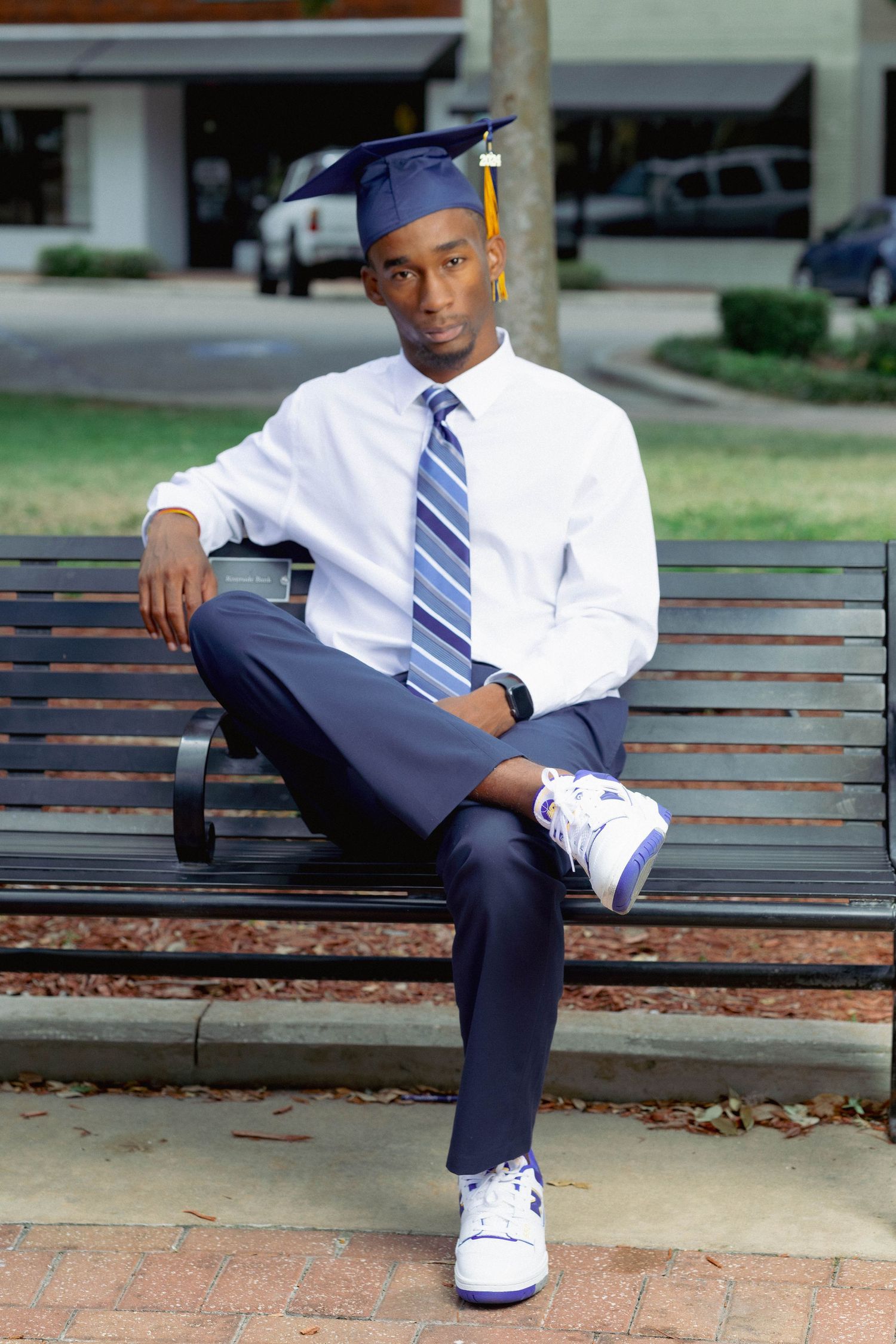 Young Black male graduate in cap and tie sitting confidently on a bench in white sneakers and navy suit.