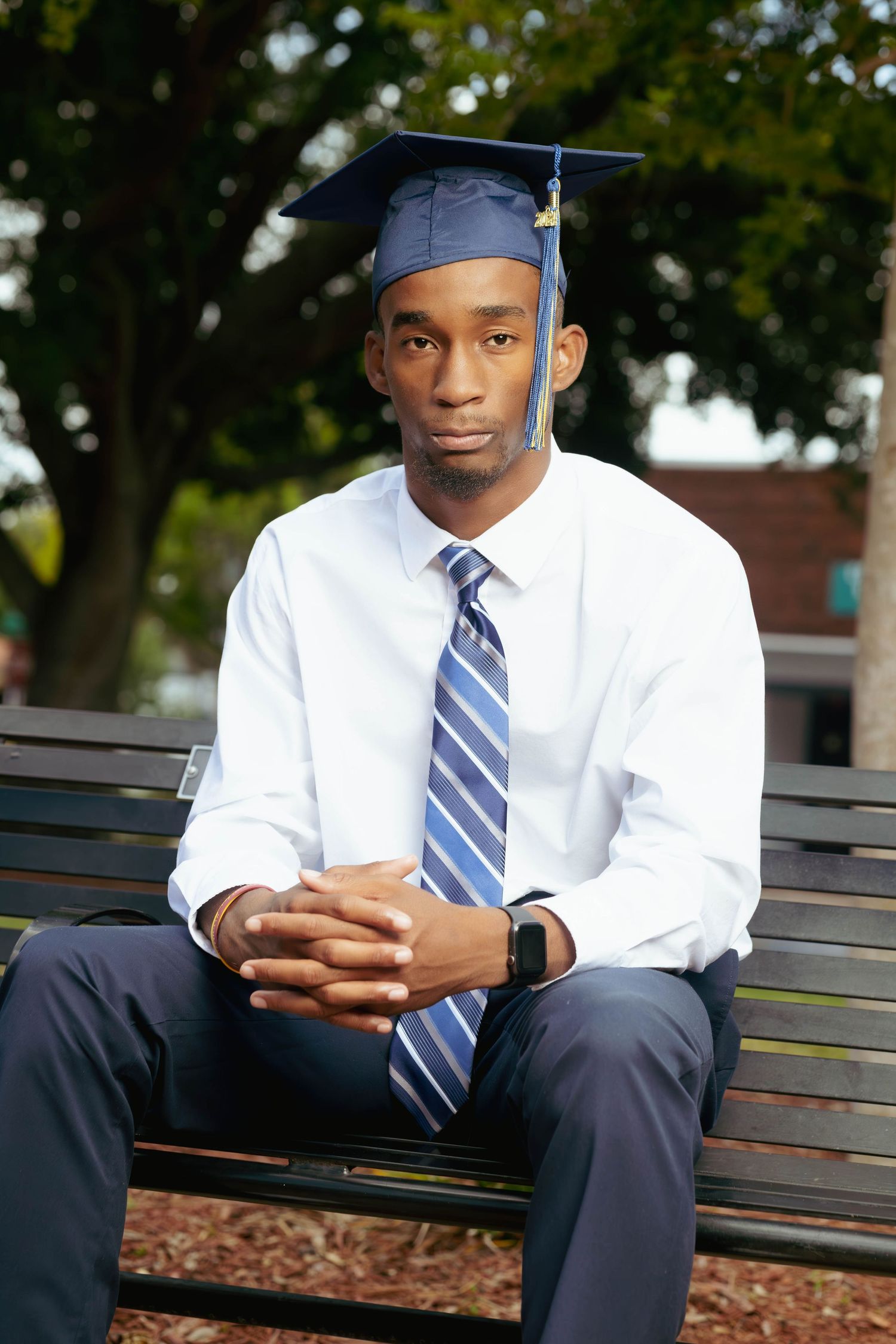 Young male graduate in cap and tie sitting on a bench outdoors for a formal graduation portrait.