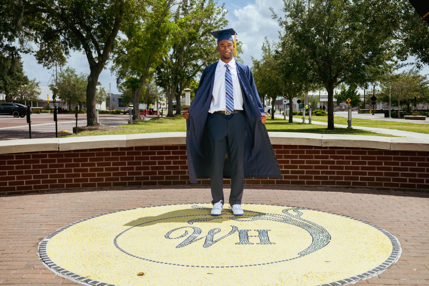 Graduate in full cap and gown standing proudly on a decorative school emblem medallion outdoors.