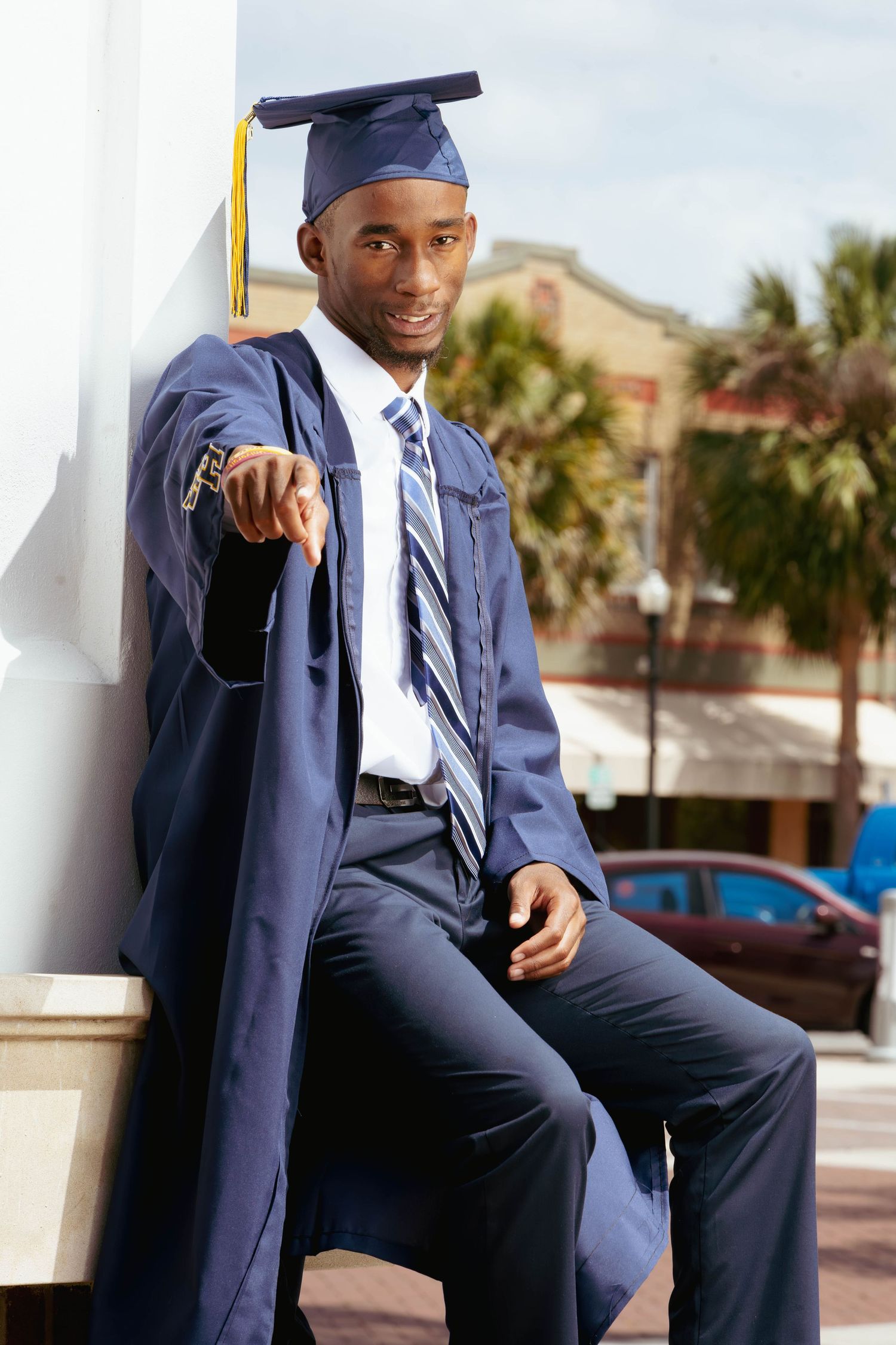 Smiling Black male graduate in navy cap and gown leaning against a white pillar outdoors.