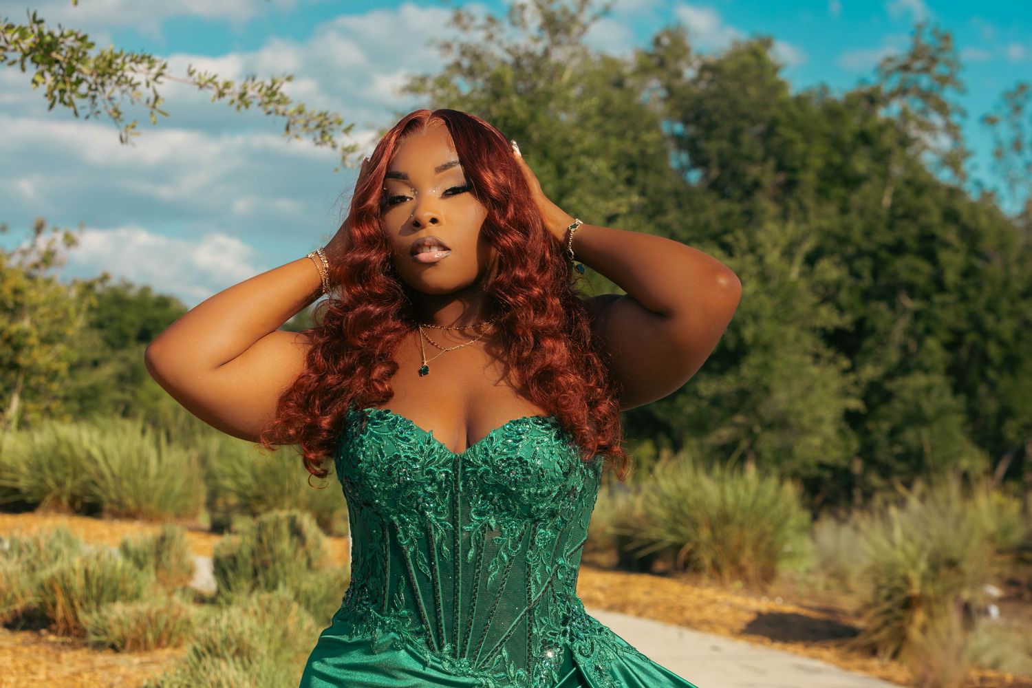 Woman with auburn curly hair in green sequin corset gown posing outdoors with hands in hair.