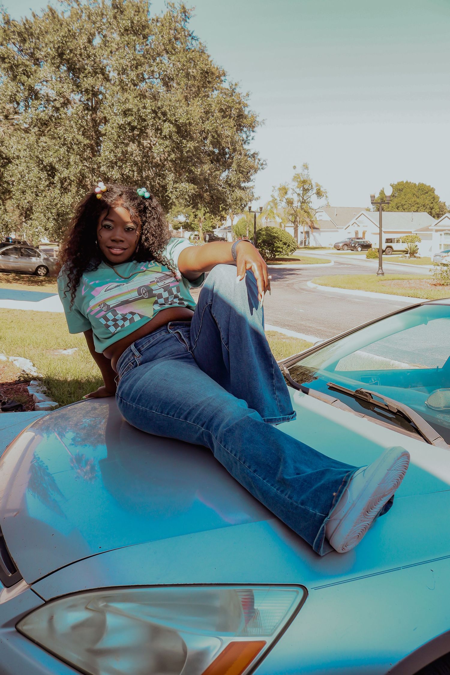 Young Black woman in teal crop top and flared jeans posing on hood of blue car outdoors.