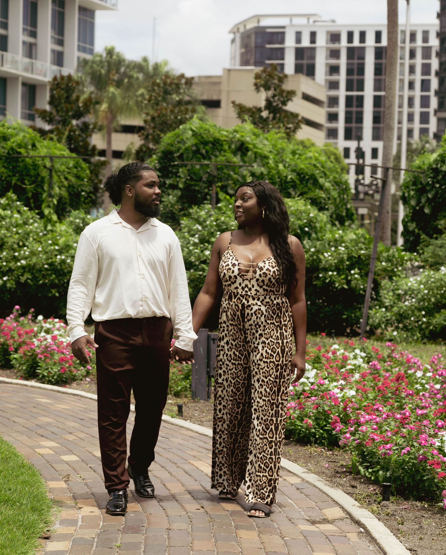 Couple in stylish outfits walking hand-in-hand through lush garden with city buildings in background.