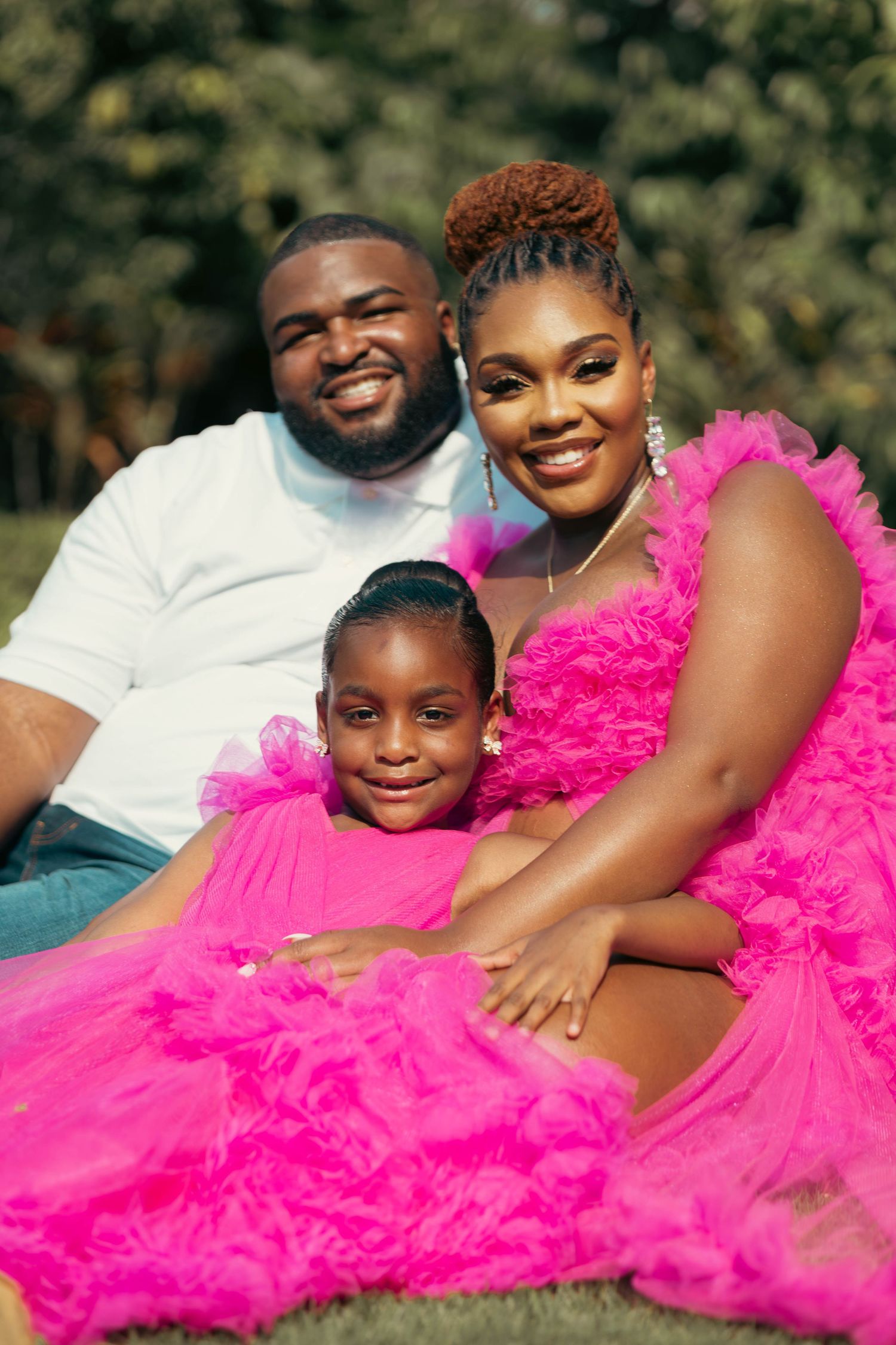 Family portrait with couple and young daughter, all wearing pink, sitting together on green grass outdoors.