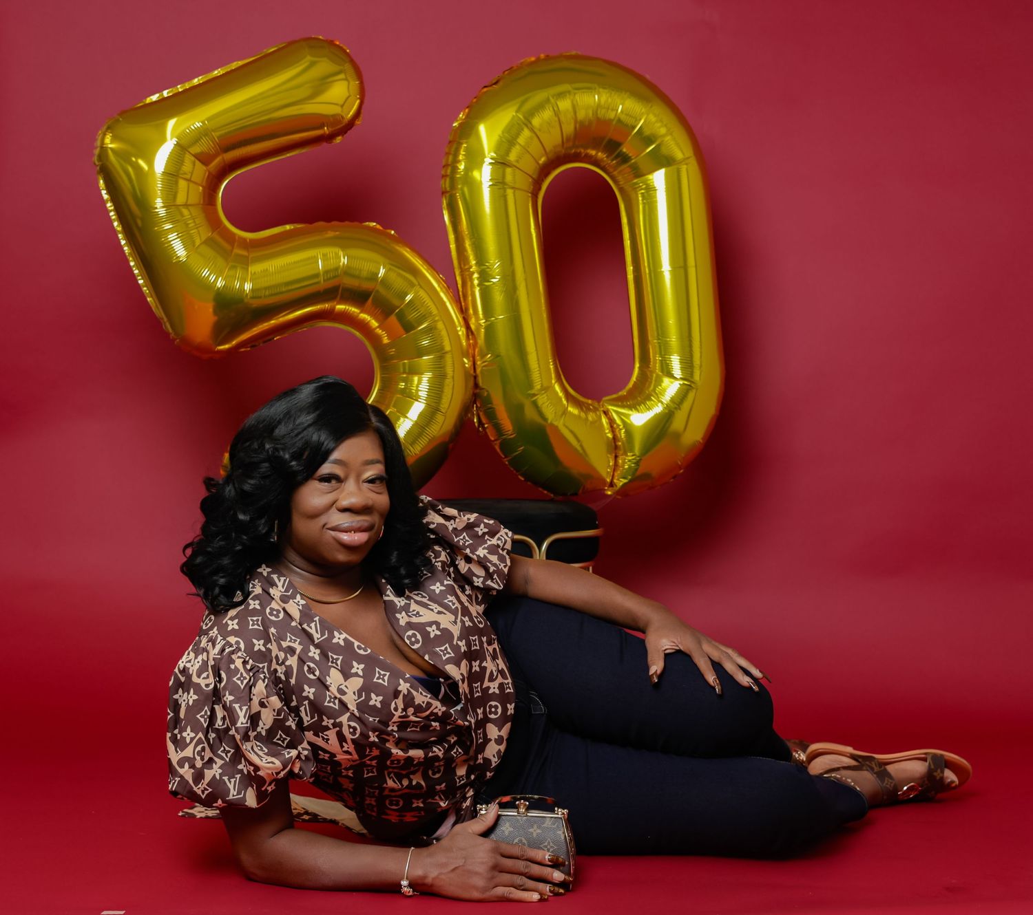 Woman celebrating 50th birthday posing with gold number balloons on red background.