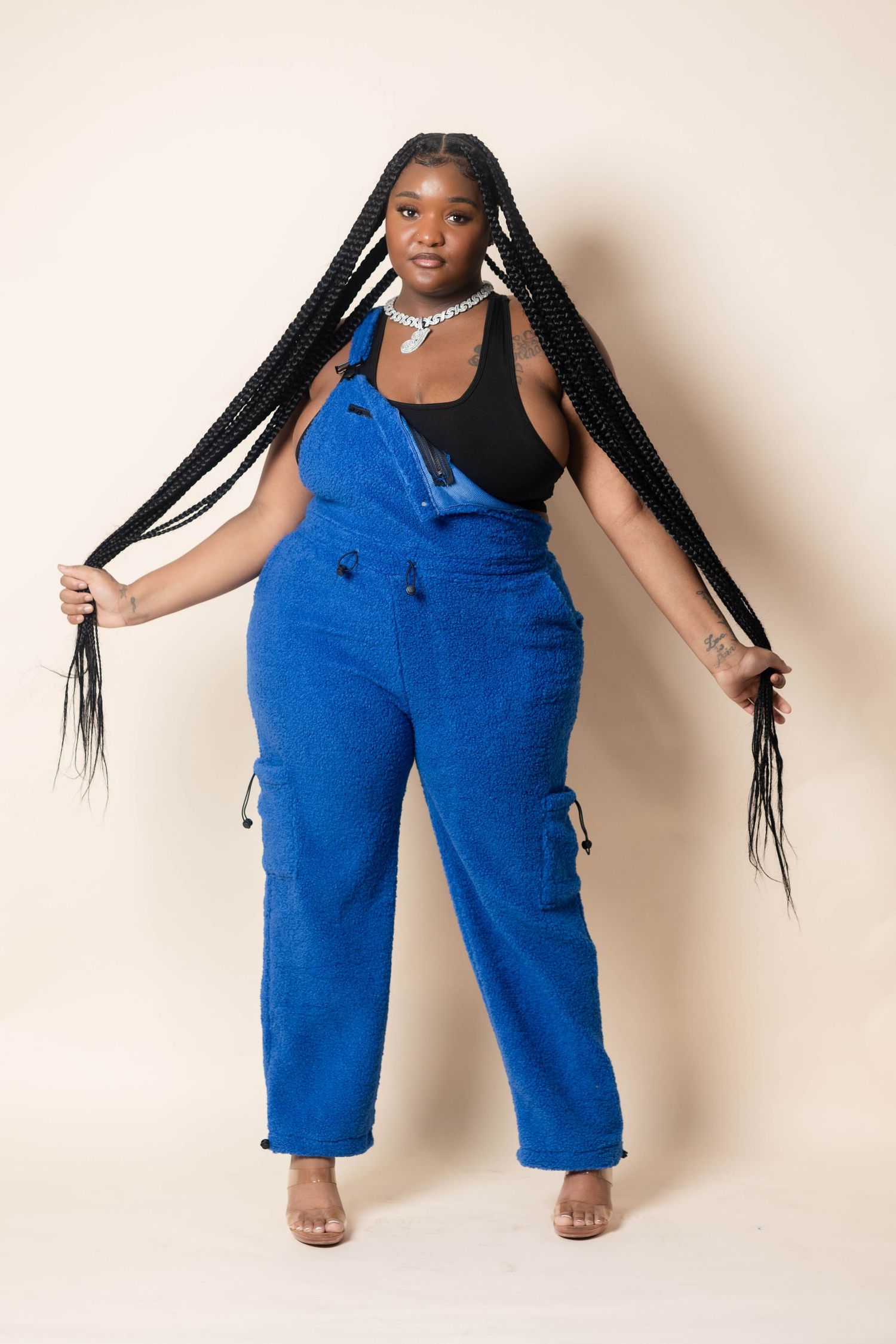 Woman in blue cargo overalls with long box braids and silver chain necklace.