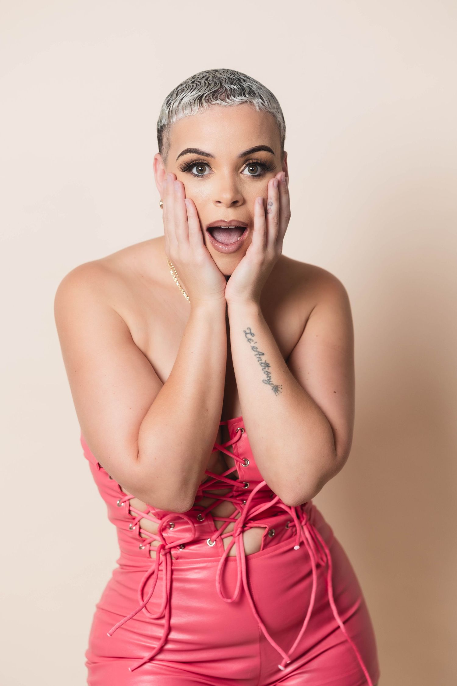 Woman with short silver hair in pink lace-up outfit with surprised expression.