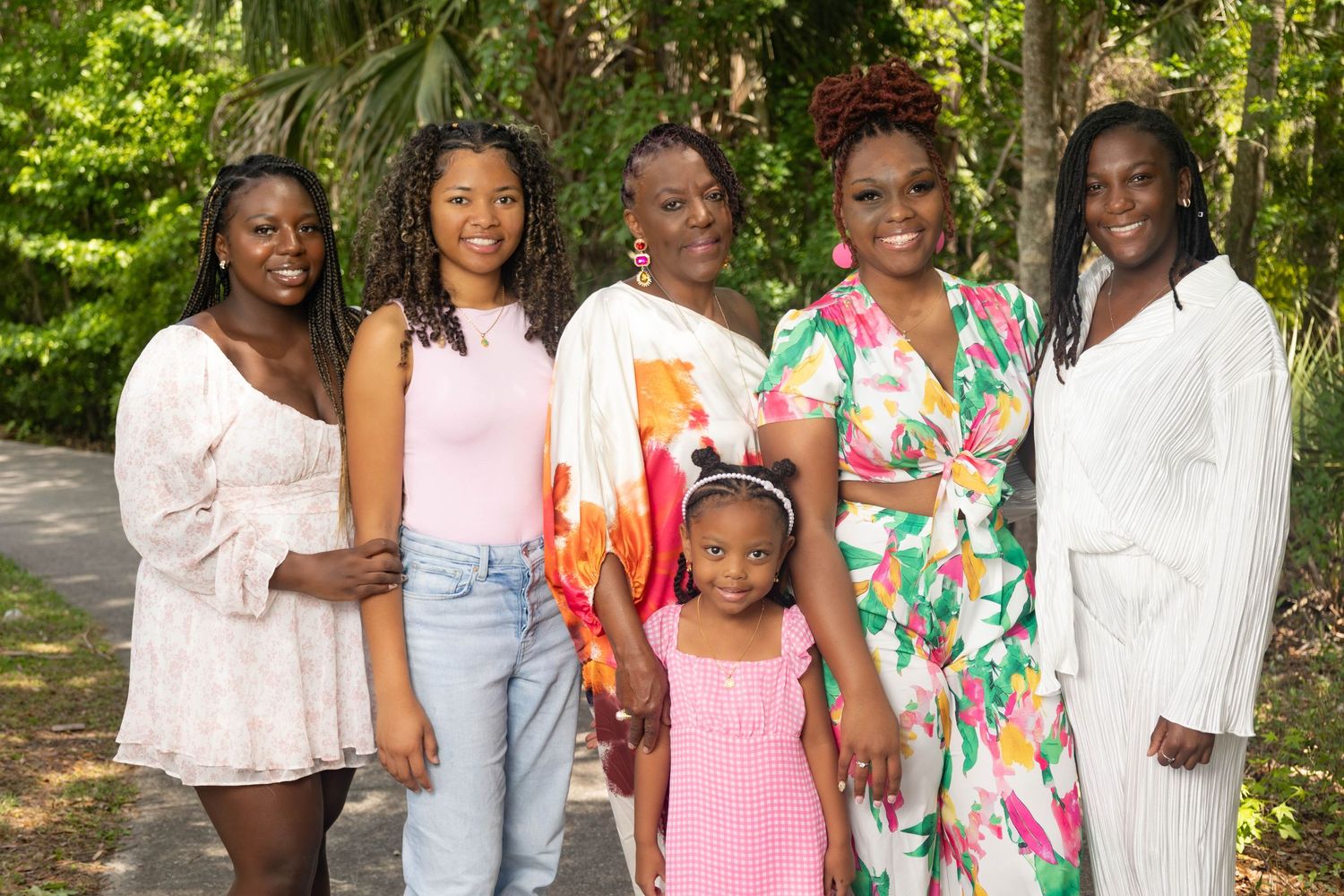 Group of six women and a young girl smiling together in a lush green outdoor setting.