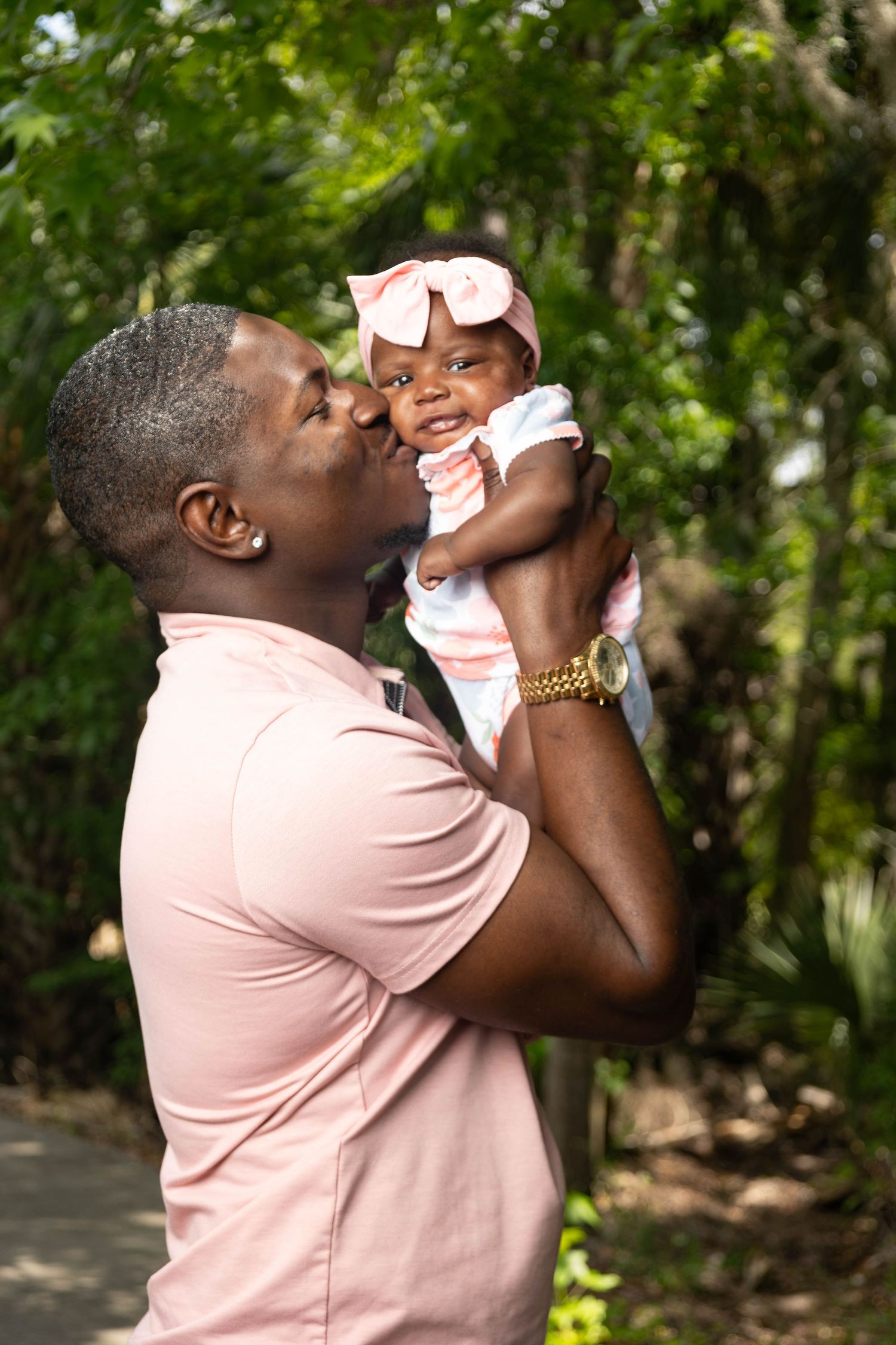 Loving father kissing baby girl with pink bow while holding her up outdoors.