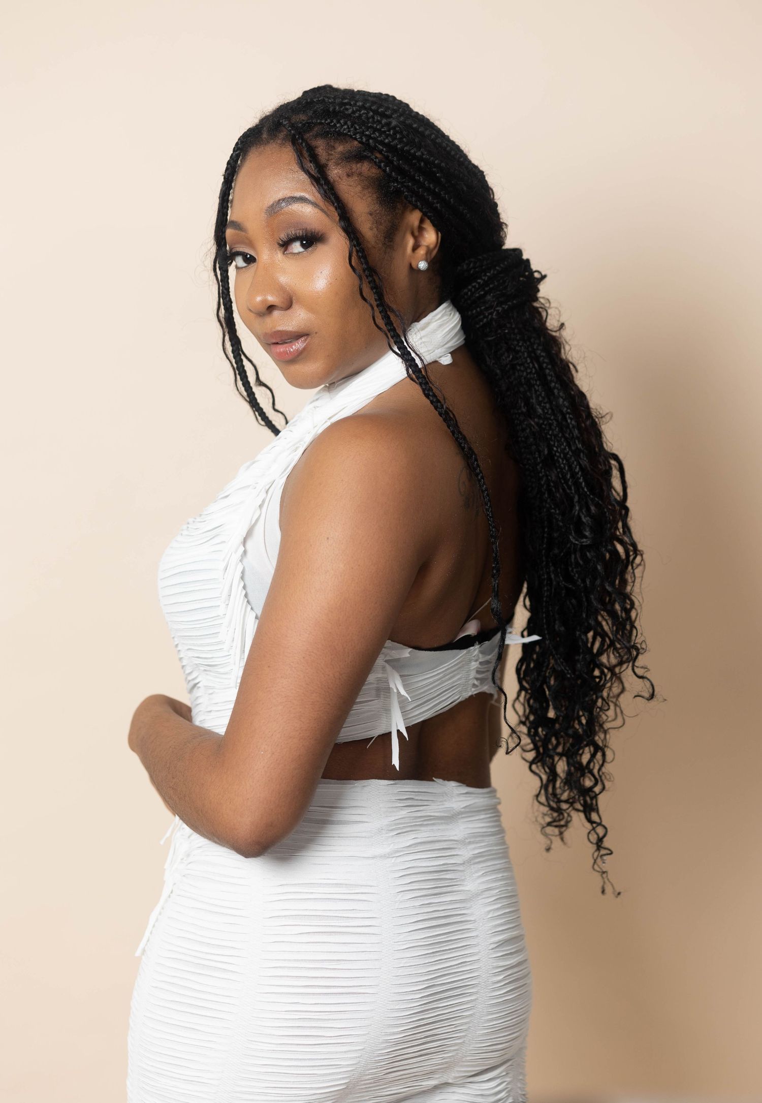 Young woman in white backless crop top and skirt with long black braids poses elegantly.