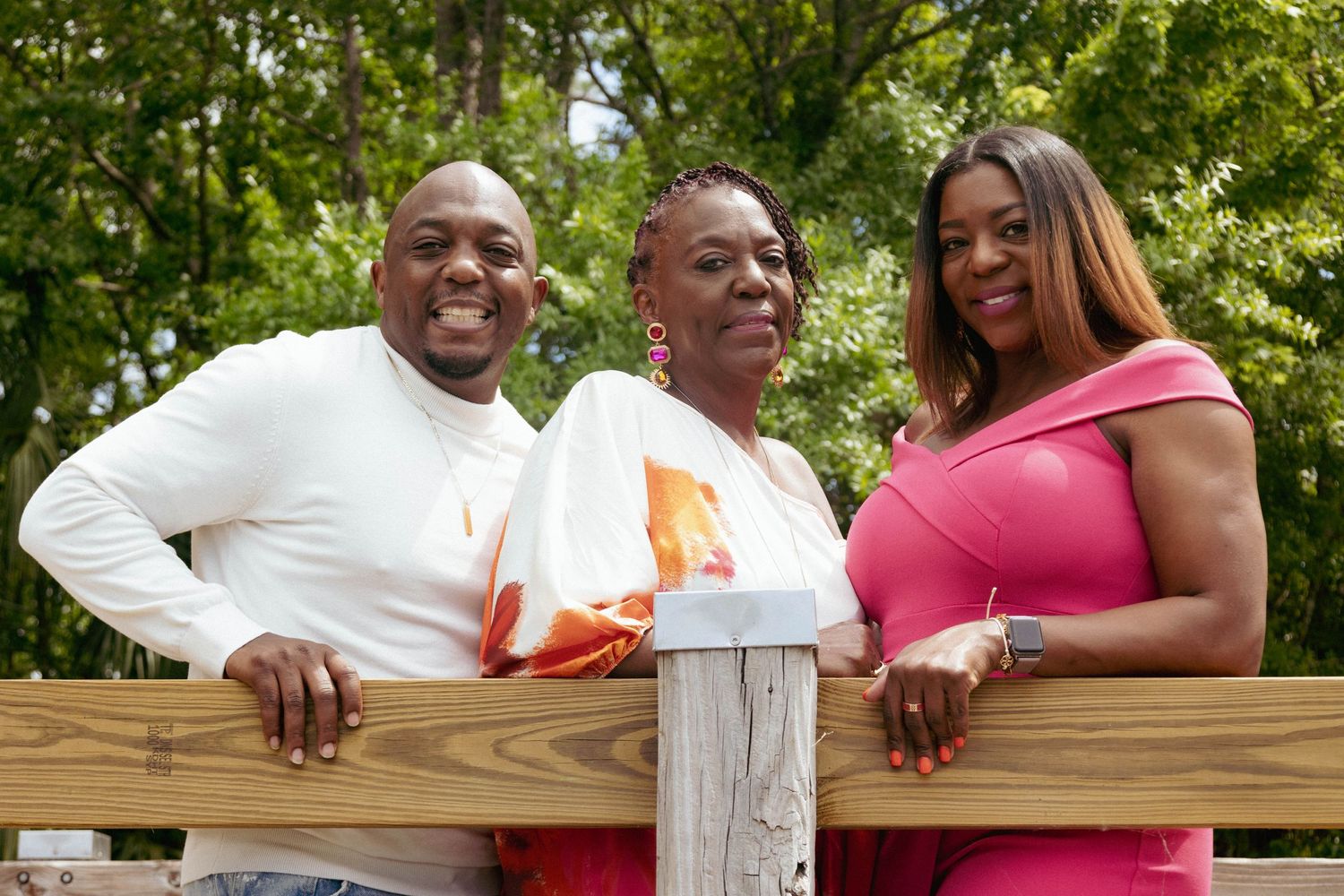 Three adults smiling and leaning on wooden fence surrounded by green trees outdoors.