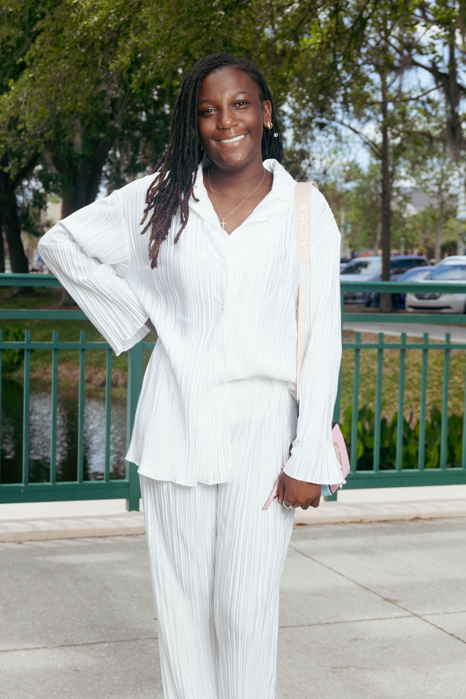 Young Black woman in white striped pantsuit smiling outdoors near green railing and trees.