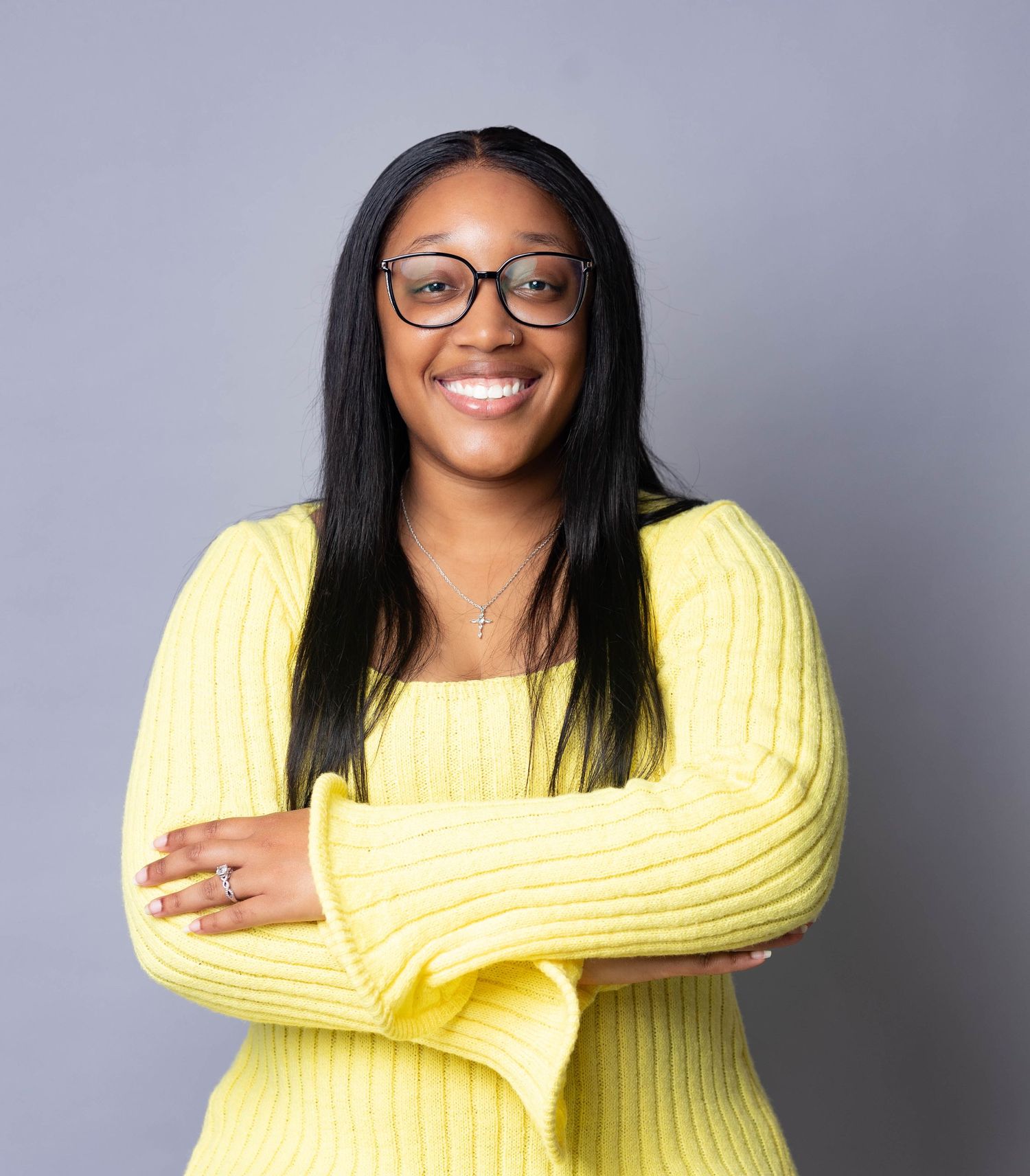 Smiling Black woman in yellow sweater with glasses posing arms crossed on gray background.