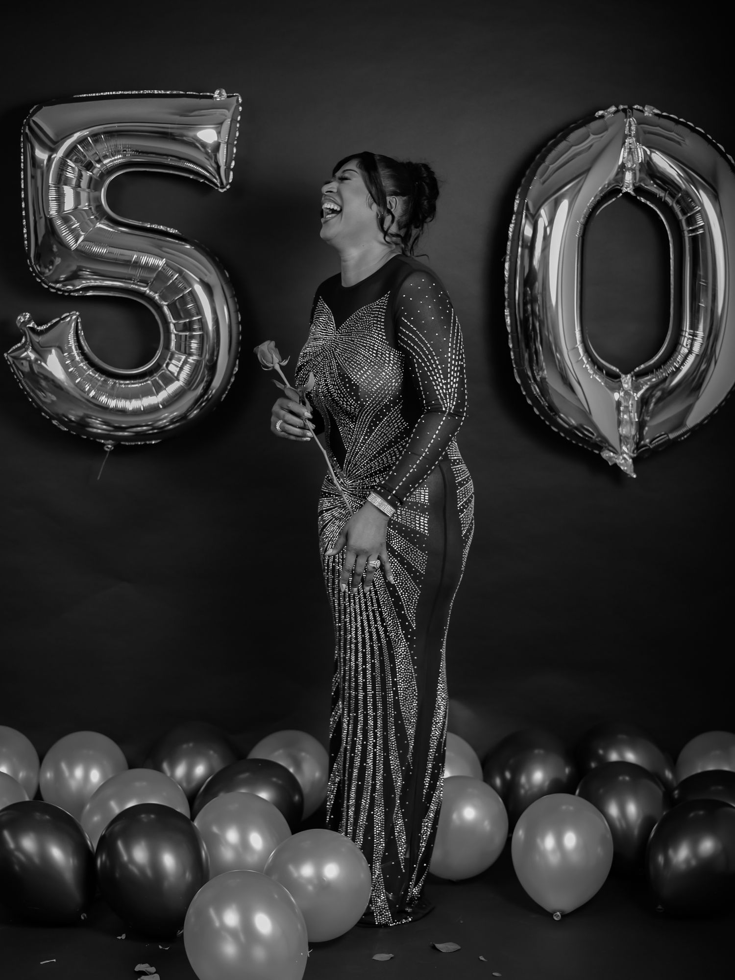 Black and white photo of woman laughing in sequin gown beside large 50 balloons at birthday celebration.