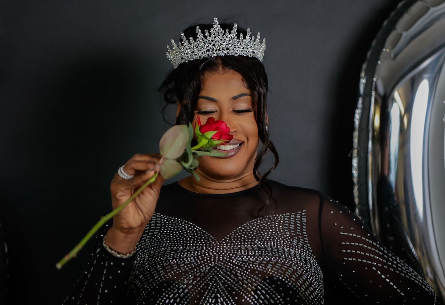 A woman wearing a diamond crown smells a red rose in a sparkly black gown.