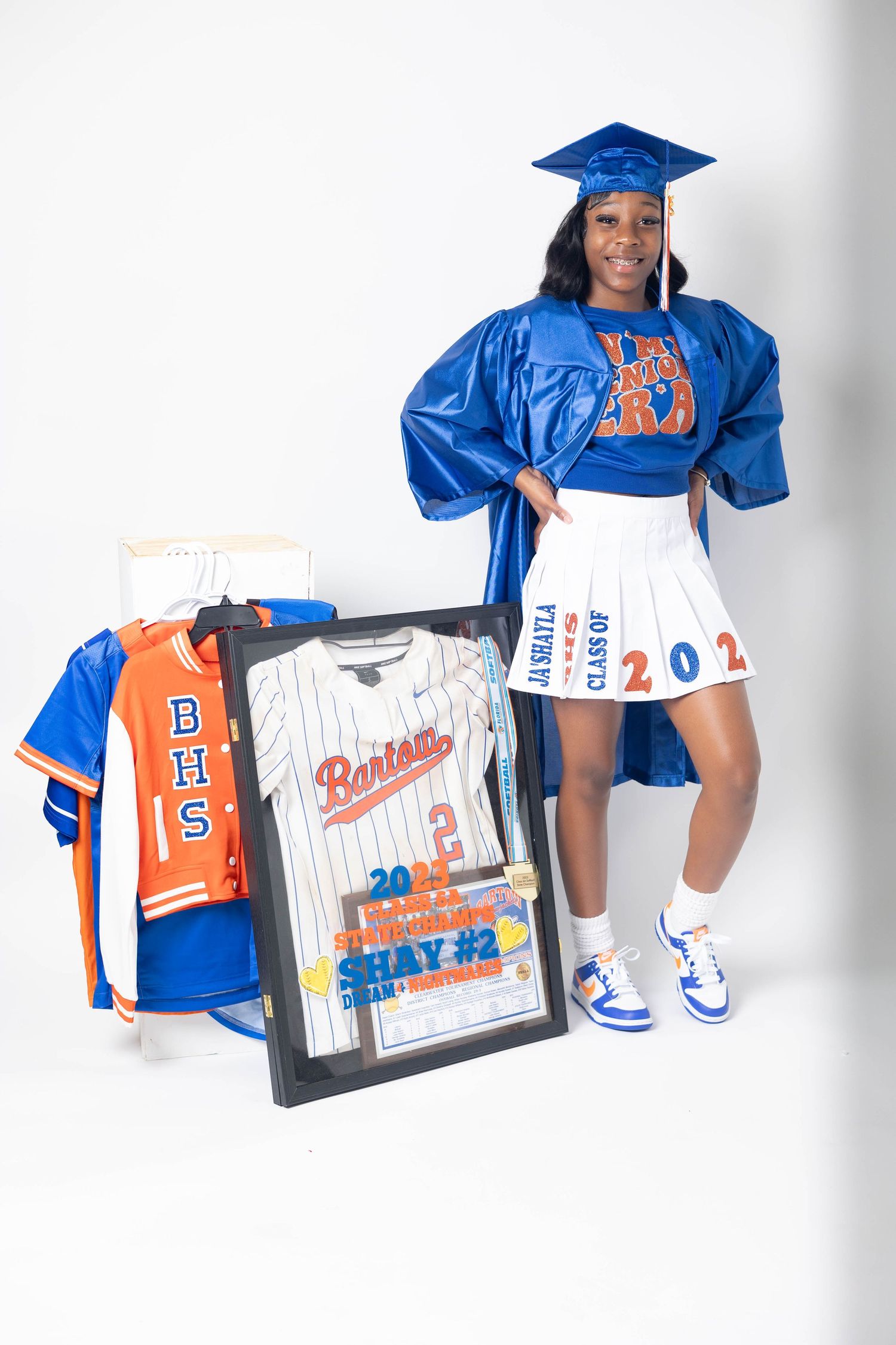 Young woman in blue graduation cap and gown poses with Bartow High School baseball jersey and framed award.