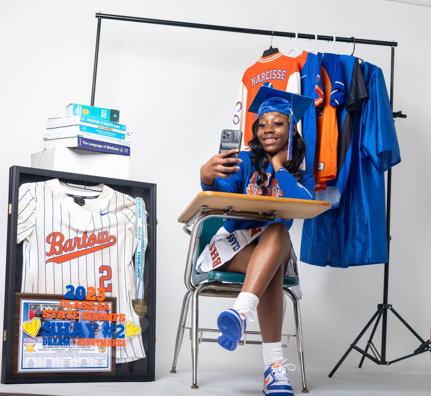 Graduate in blue cap sits at desk with framed Bartow jersey, phone, and sports uniforms on rack behind her.