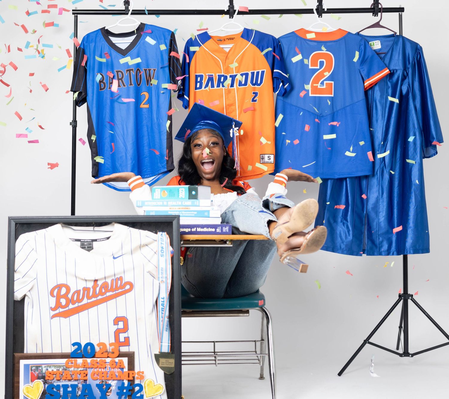 Smiling graduate surrounded by colorful confetti and multiple Bartow baseball jerseys displayed on rack.