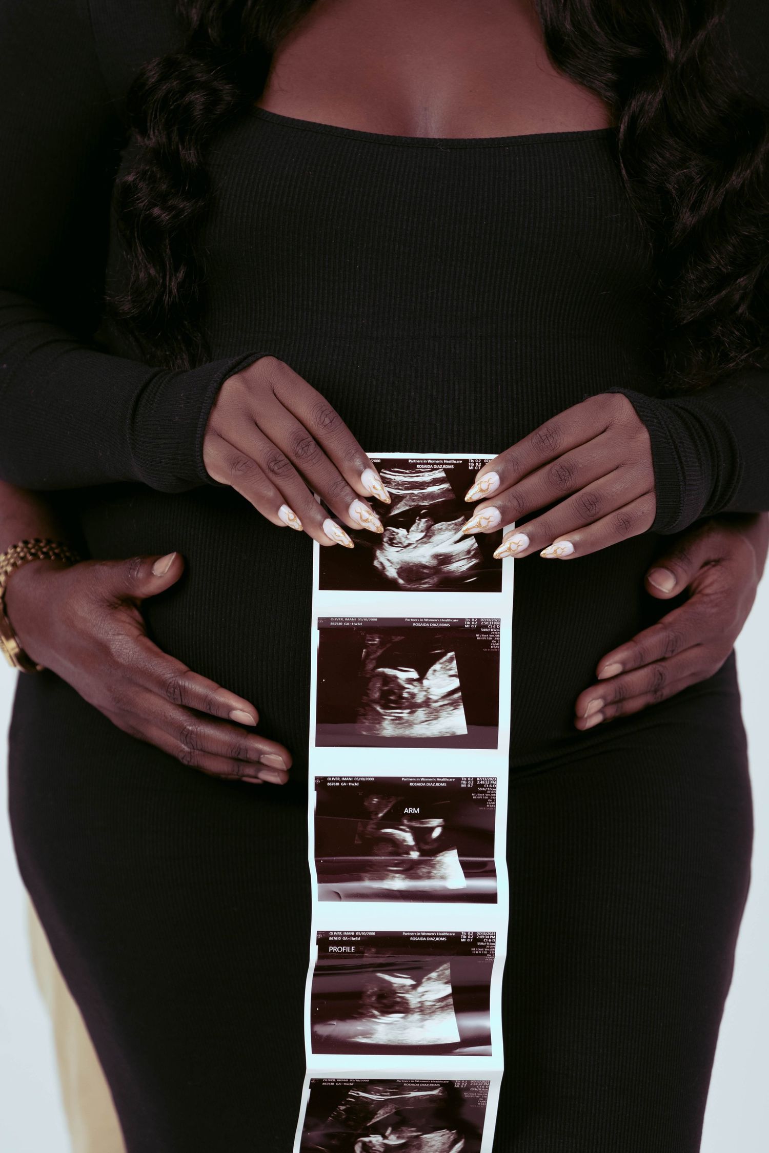 Pregnant woman in black dress holds ultrasound strip photos against her baby bump.