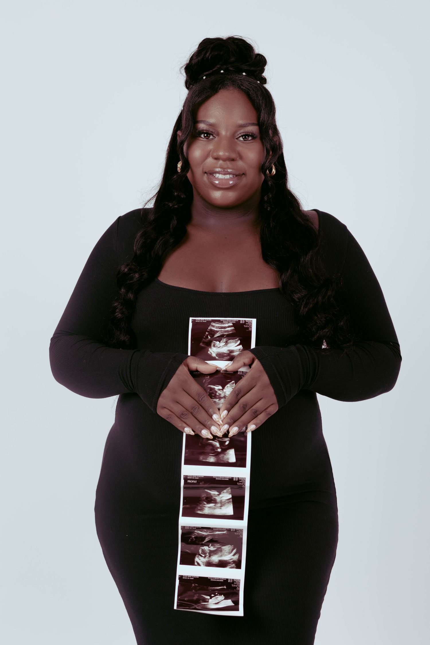 Smiling pregnant woman in black dress holds ultrasound strip photos while posing for maternity portrait.