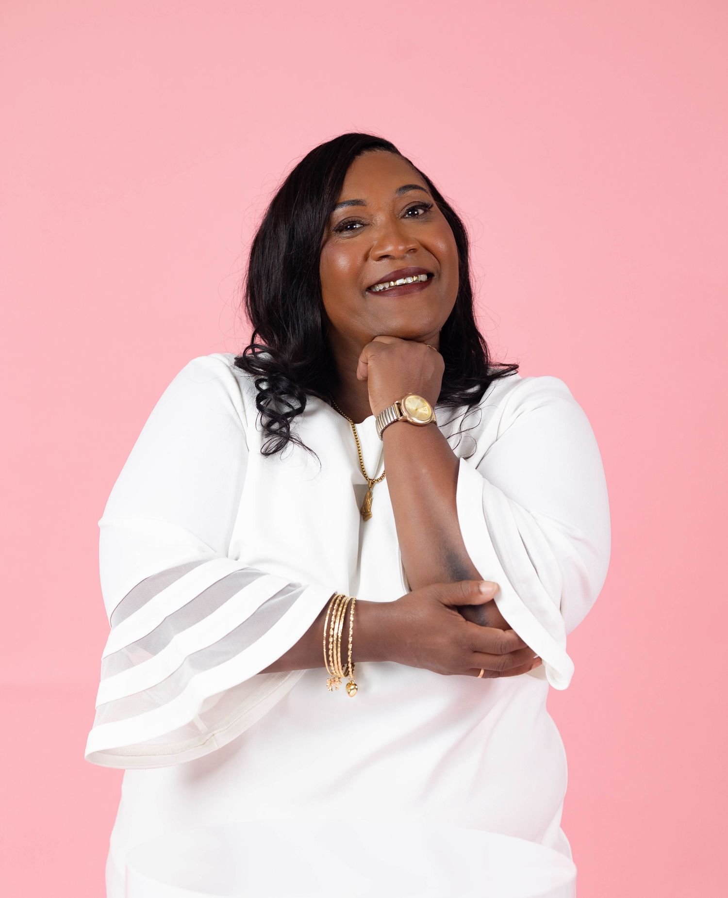 Woman in white dress smiles warmly against a pink background, wearing gold jewelry and bracelets.