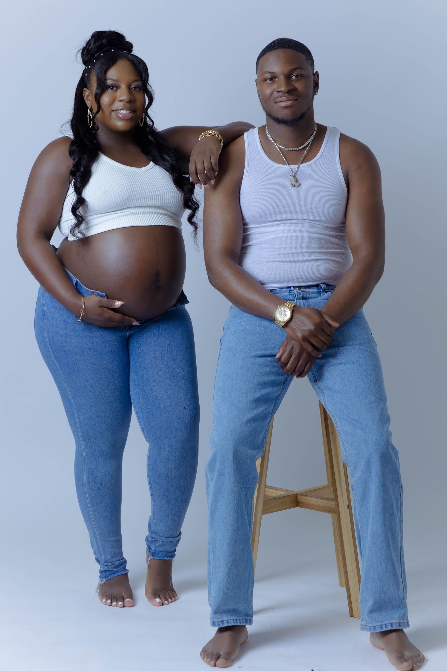 Expecting couple in matching white tanks and jeans pose together for maternity photo session.