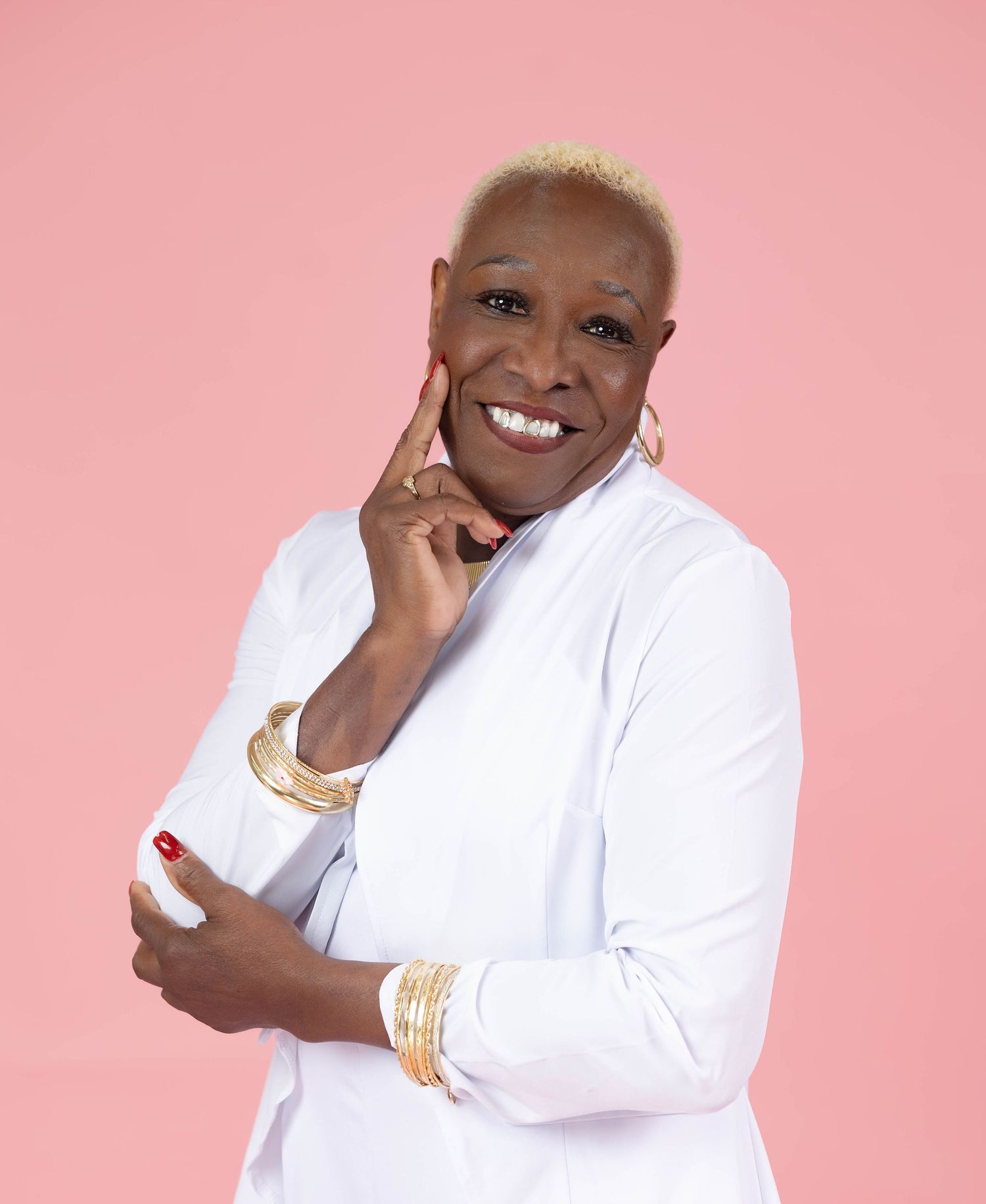 Stylish woman with blonde hair in white outfit poses confidently with gold jewelry on pink background.